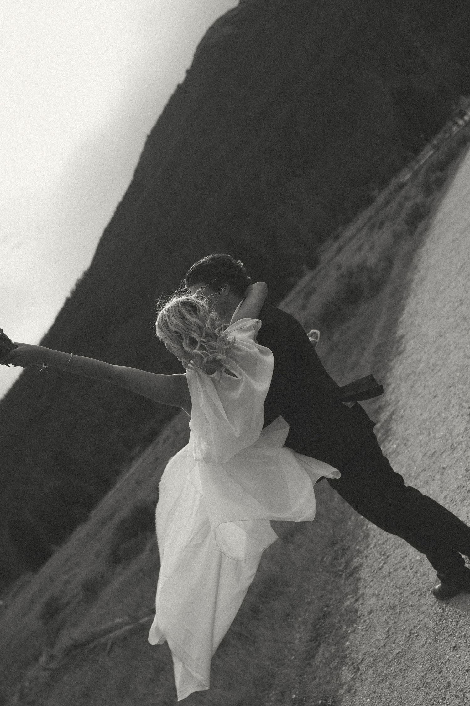 Black and white photo of a bride in a flowing white gown kissing her groom on a mountain trail with her arm outstretched.