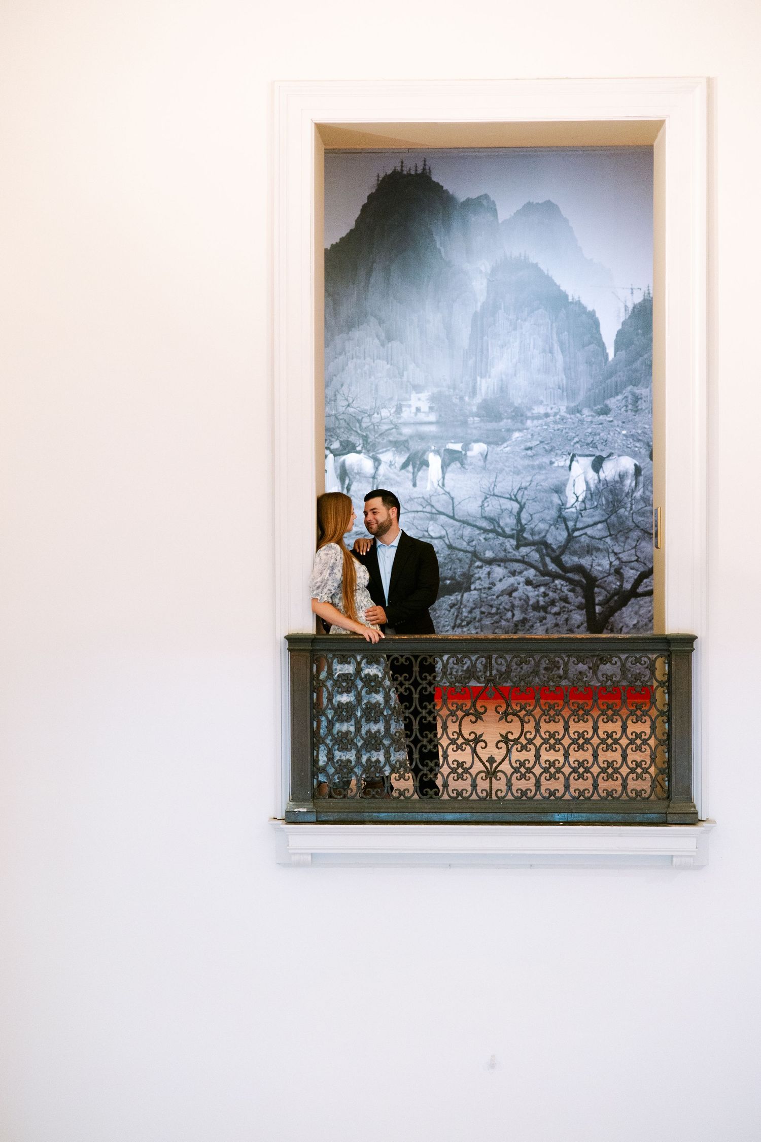 Engaged couple standing together on a small ornate balcony in front of a large black and white mountain landscape artwork.