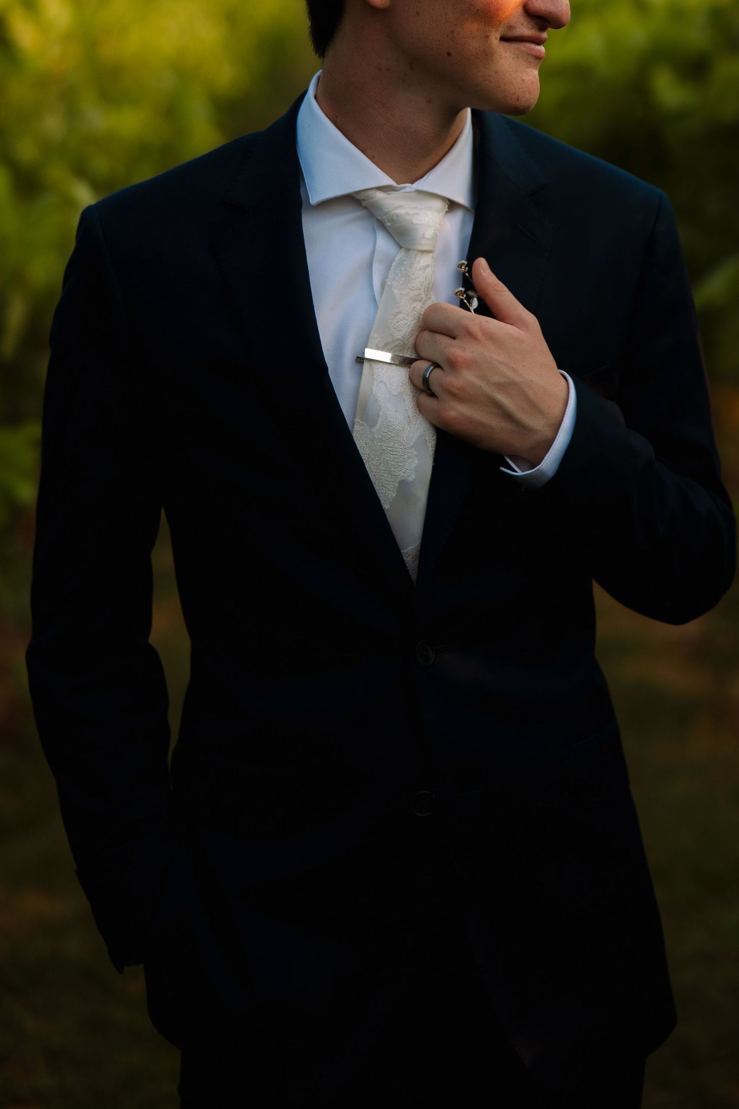 Groom in a navy suit with a patterned tie and silver tie bar adjusts his lapel in warm golden evening light.