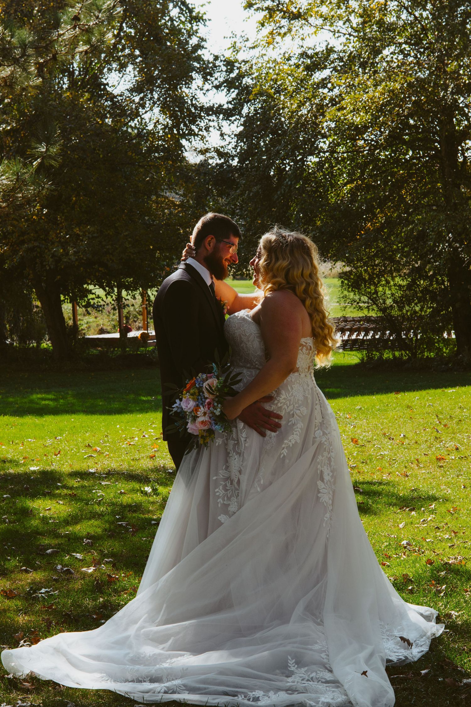 Bride in a flowing lace gown holding a bouquet gazes at her bearded groom in a dark suit on a sunny, tree-lined lawn.