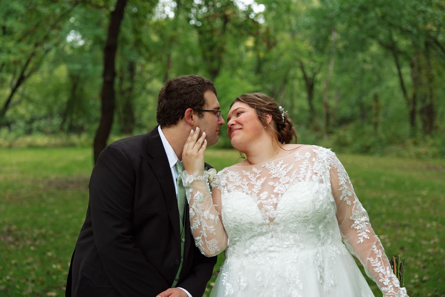 Bride in a white lace wedding gown and groom in a dark suit touch noses affectionately while posing outdoors in a green, tree-filled park.