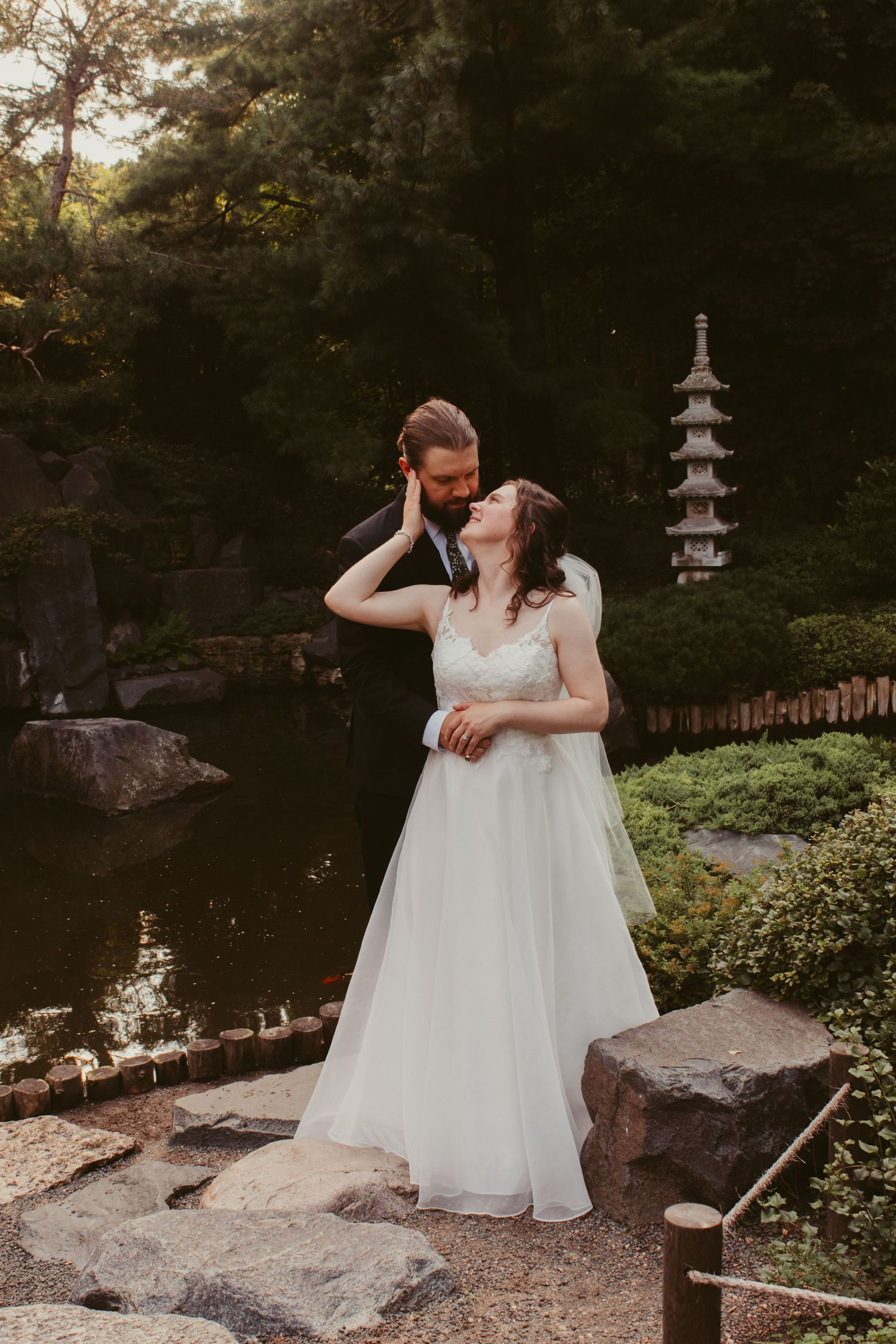 Groom embraces his bride beside a tranquil pond in a Japanese garden with a stone pagoda lantern in the background.