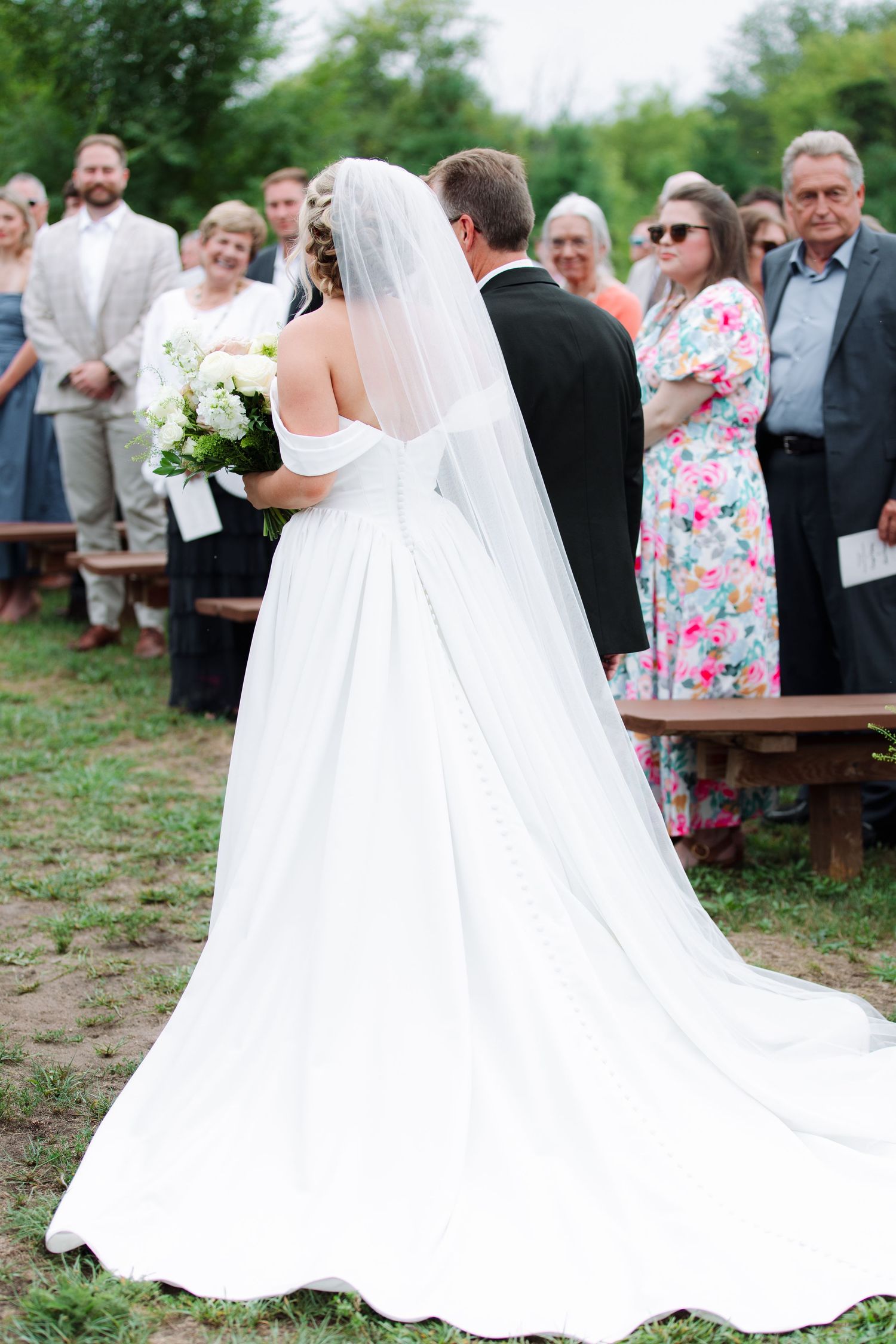 Bride in a white off-the-shoulder gown with a long veil walks down the aisle at an outdoor wedding as guests look on.