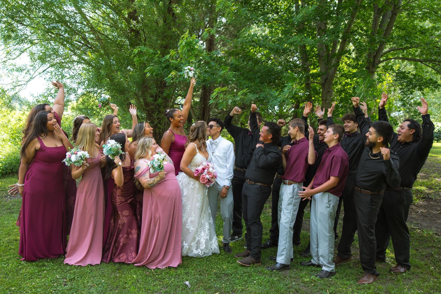 Joyful wedding party raises their arms in celebration around the bride and groom, with bridesmaids holding bouquets under tall green trees.