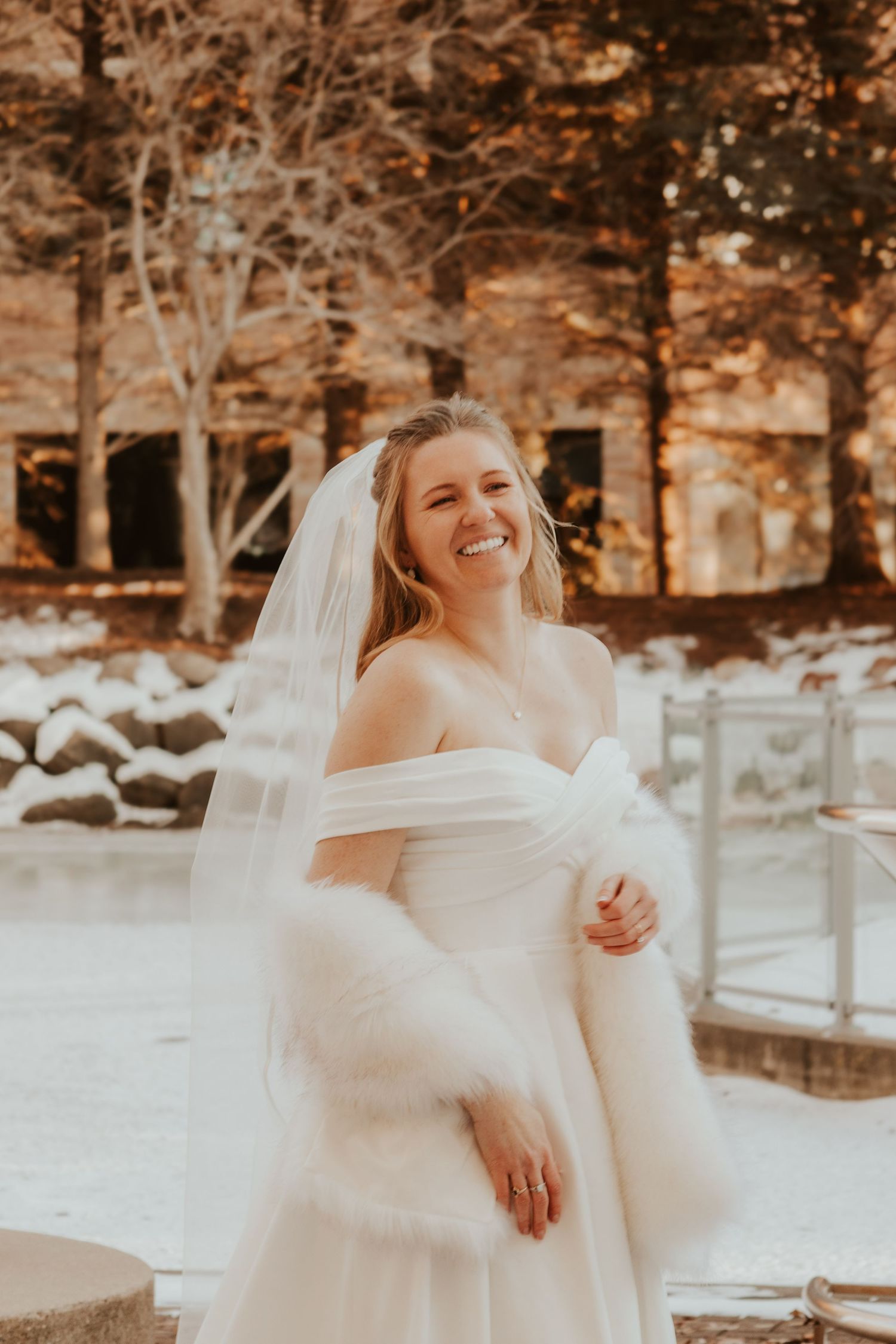 Smiling bride in an off-shoulder white wedding gown, long veil and faux fur wrap stands outdoors in a snowy winter setting.