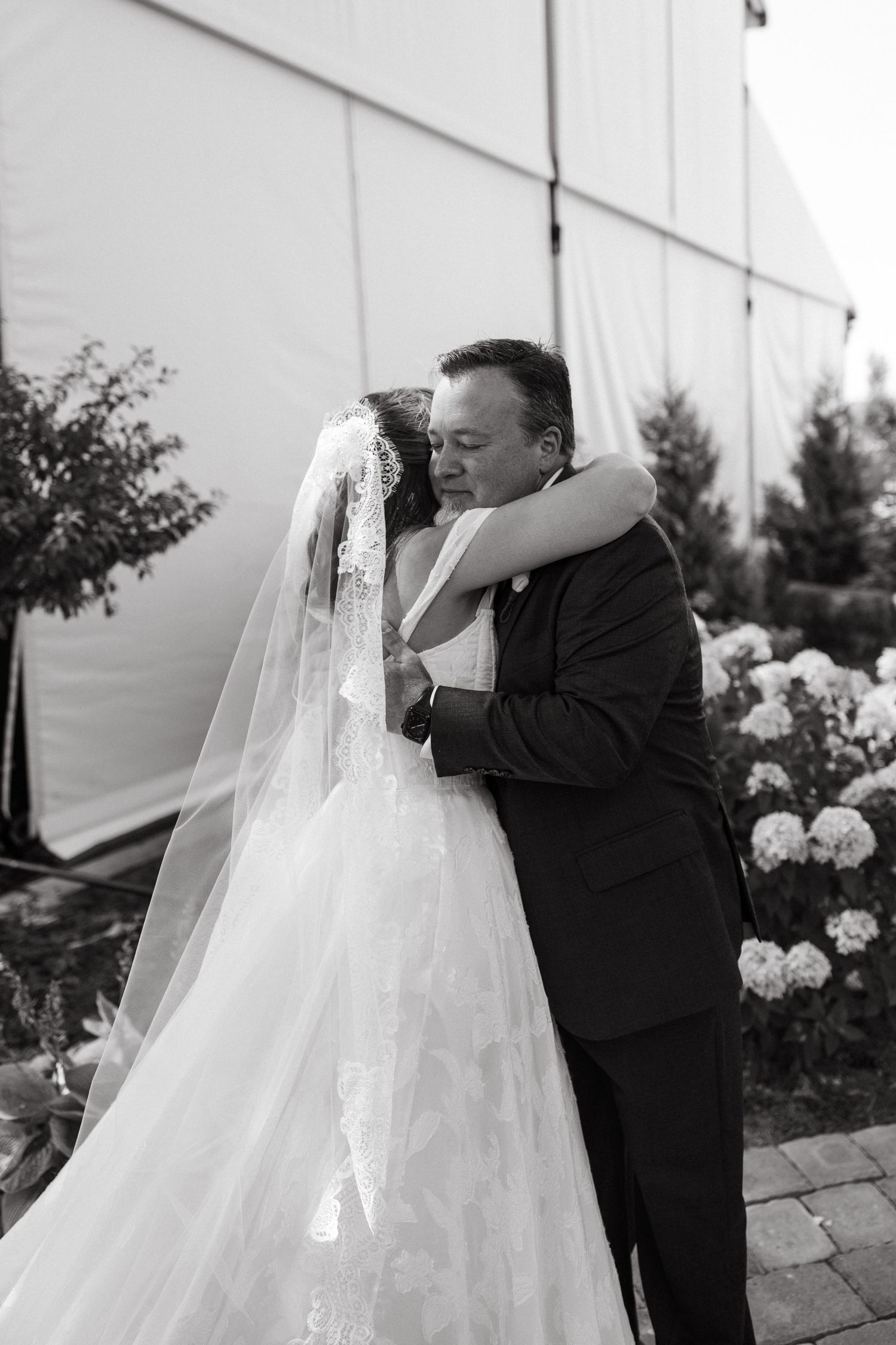 Black and white photo of a father tenderly hugging his daughter, the bride, in her lace veil beside white hydrangeas.