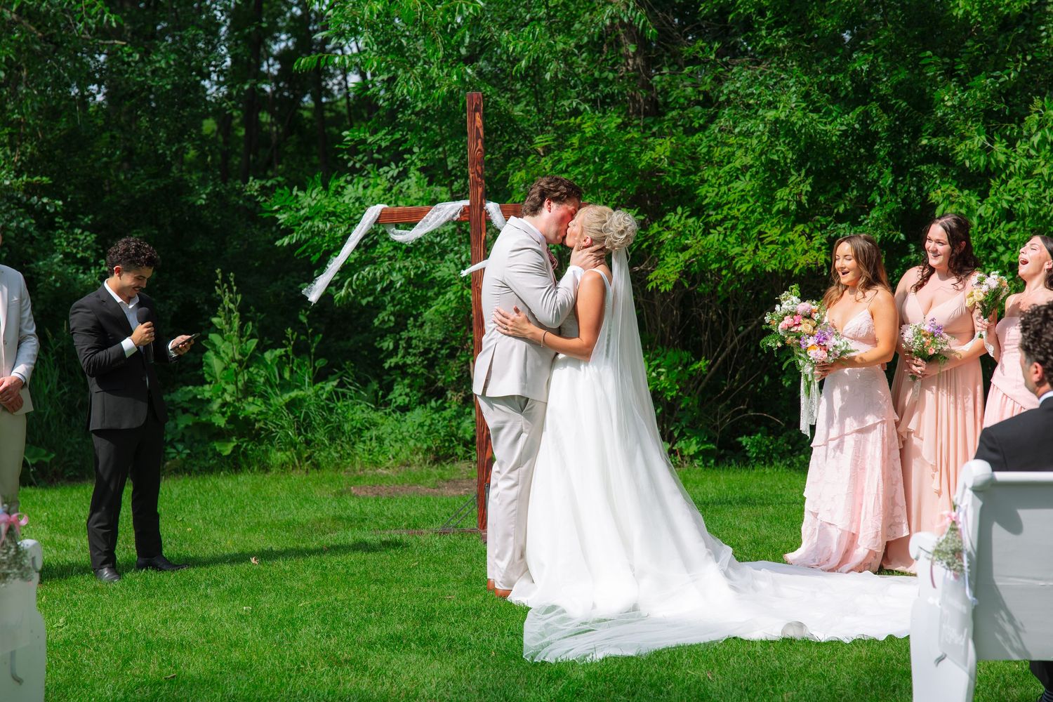 Bride and groom share a kiss at an outdoor wedding ceremony near a wooden cross as bridesmaids and guests look on.