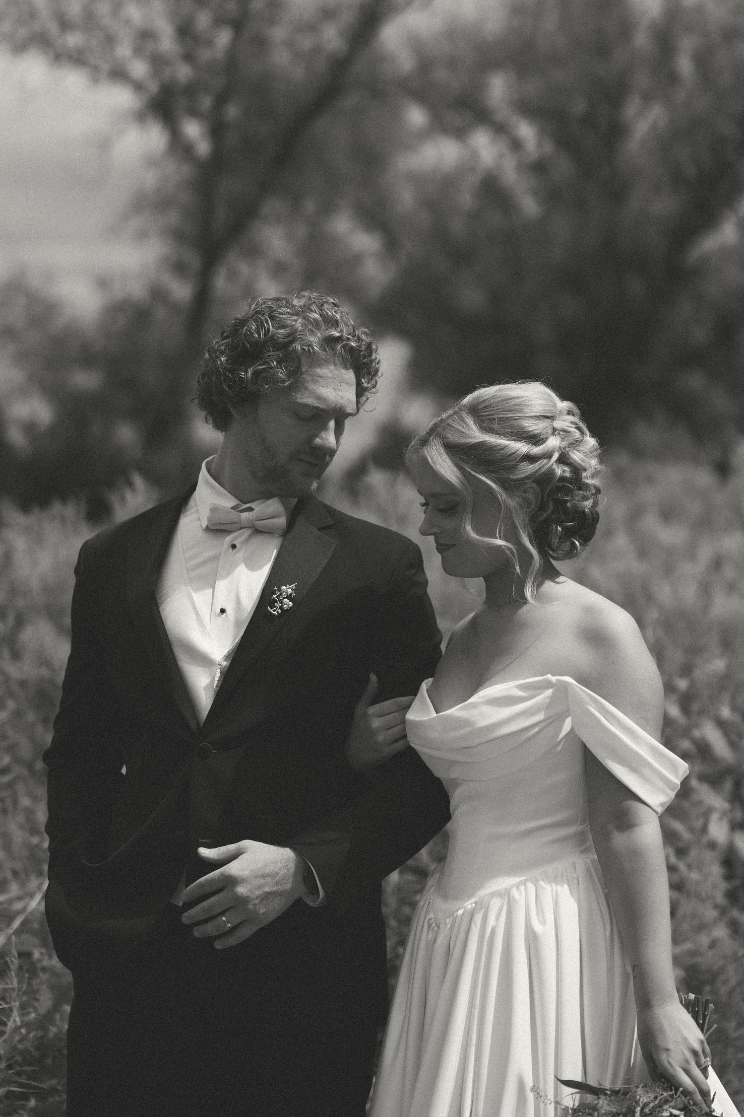 Black and white photo of a bride in an off-shoulder gown and a groom in a tuxedo with bow tie standing close in a natural outdoor field.