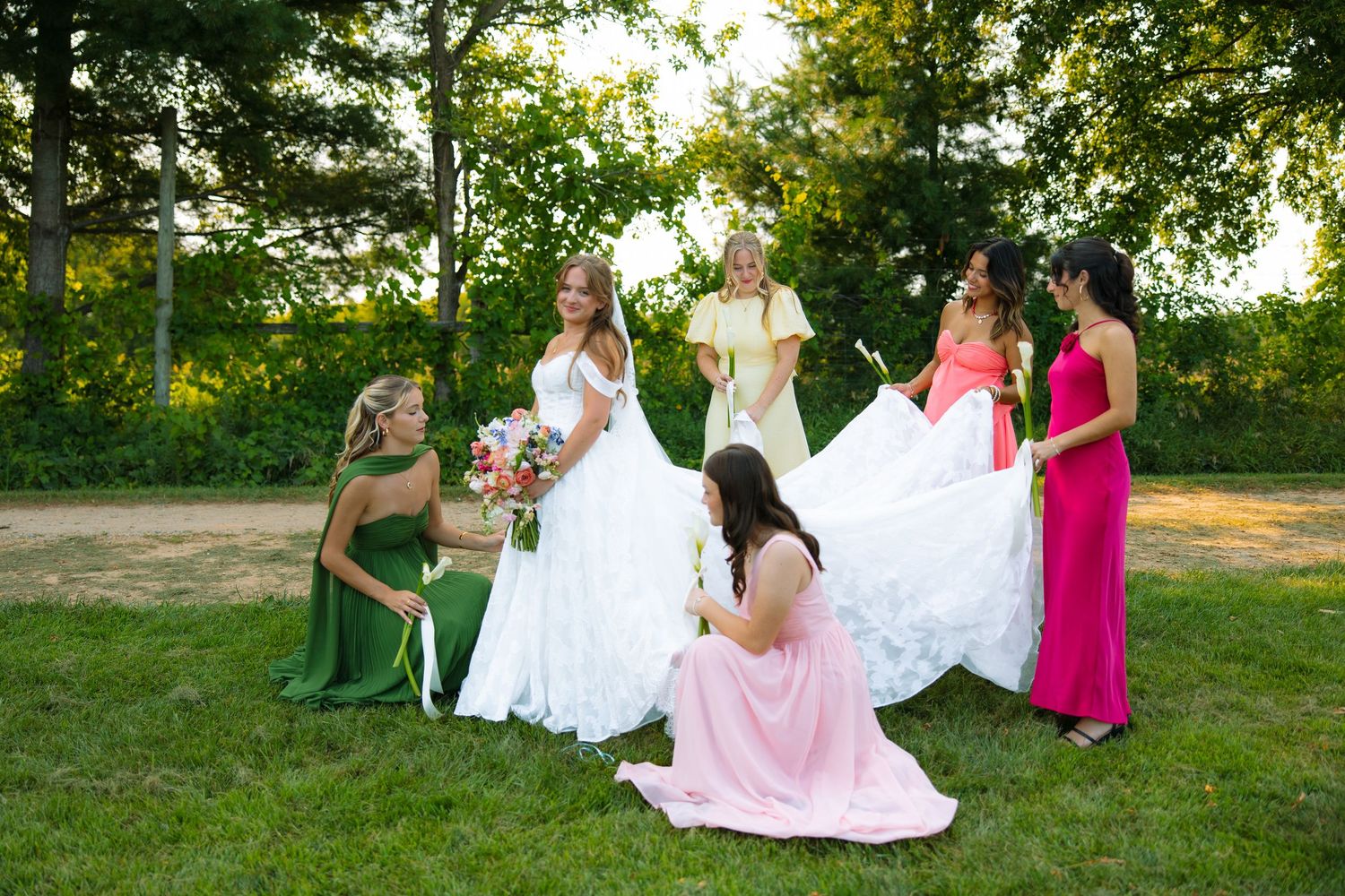 A bride in a white gown holds a bouquet while bridesmaids in green, pink, and yellow dresses gather around her on the grass.