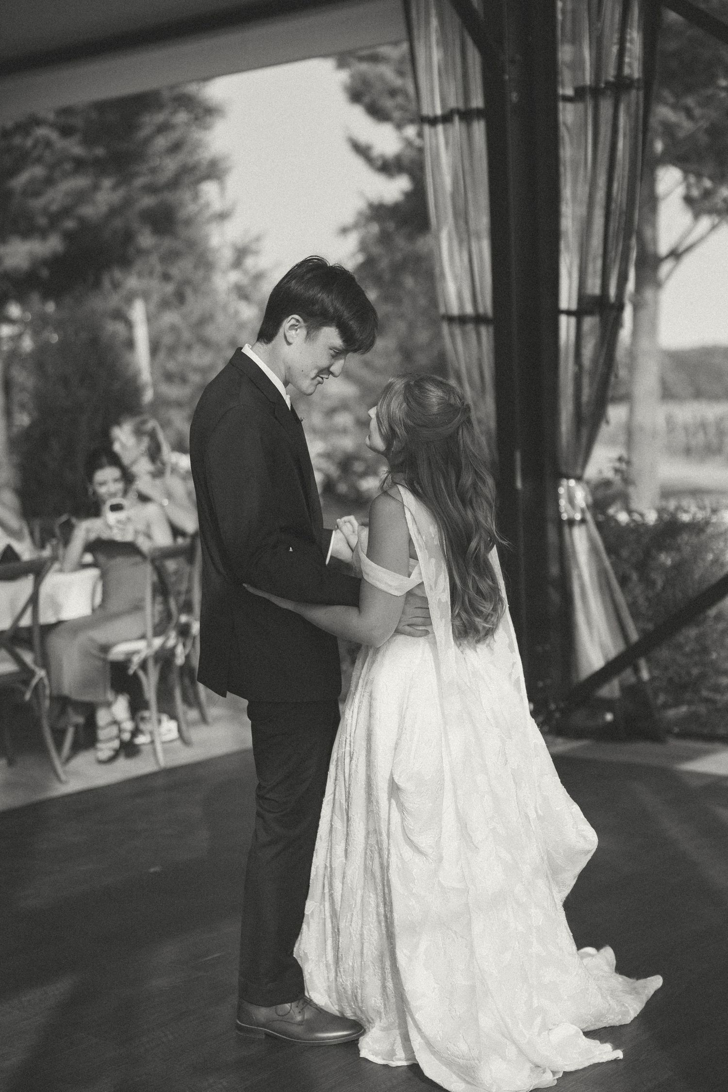 Black and white photo of a bride and groom sharing a first dance on a wooden floor under a tent as guests watch.