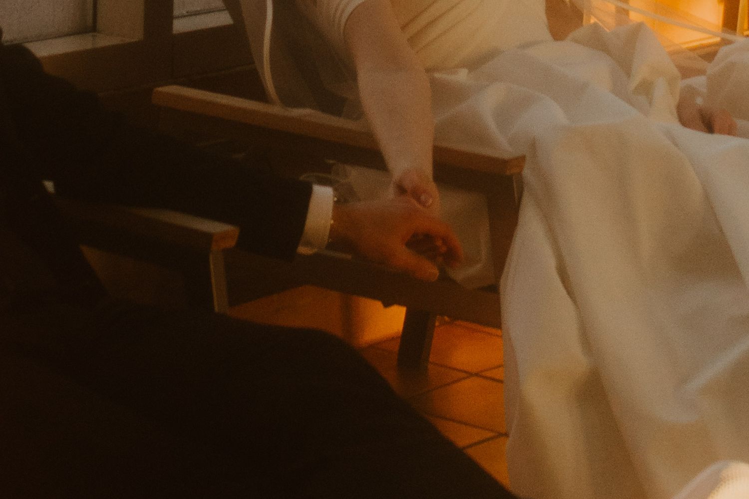 Close-up of a groom's hand holding the bride's hand as they sit together on lounge chairs in warm, amber-toned light.