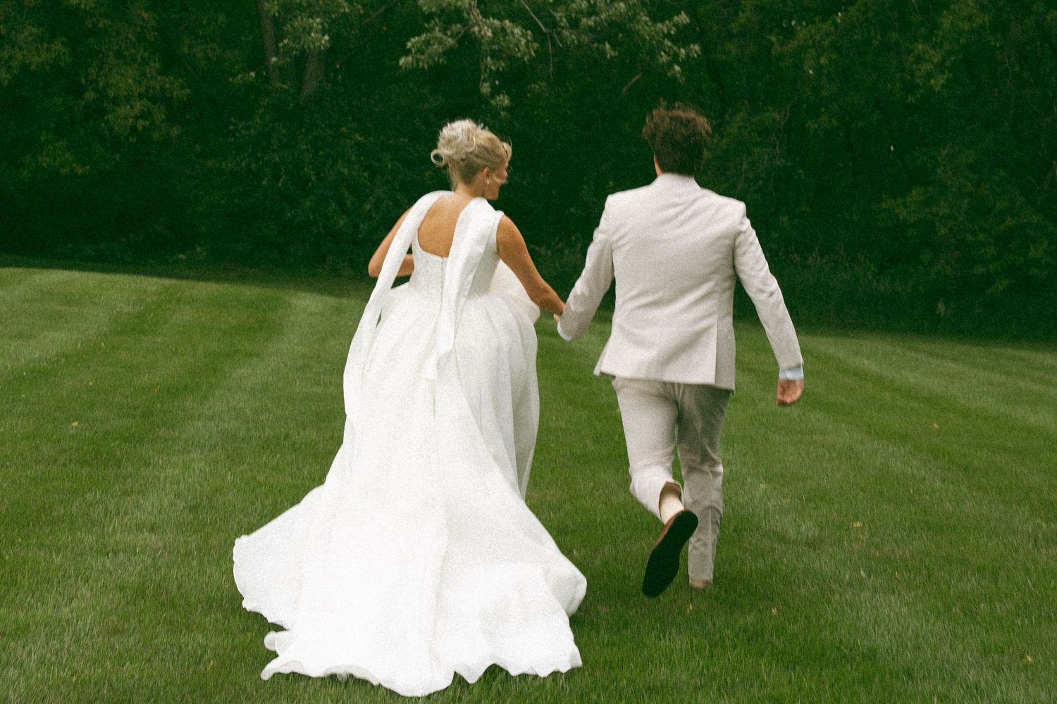 A bride in a long white gown and a groom in a beige suit hold hands and run across a green lawn toward the trees.