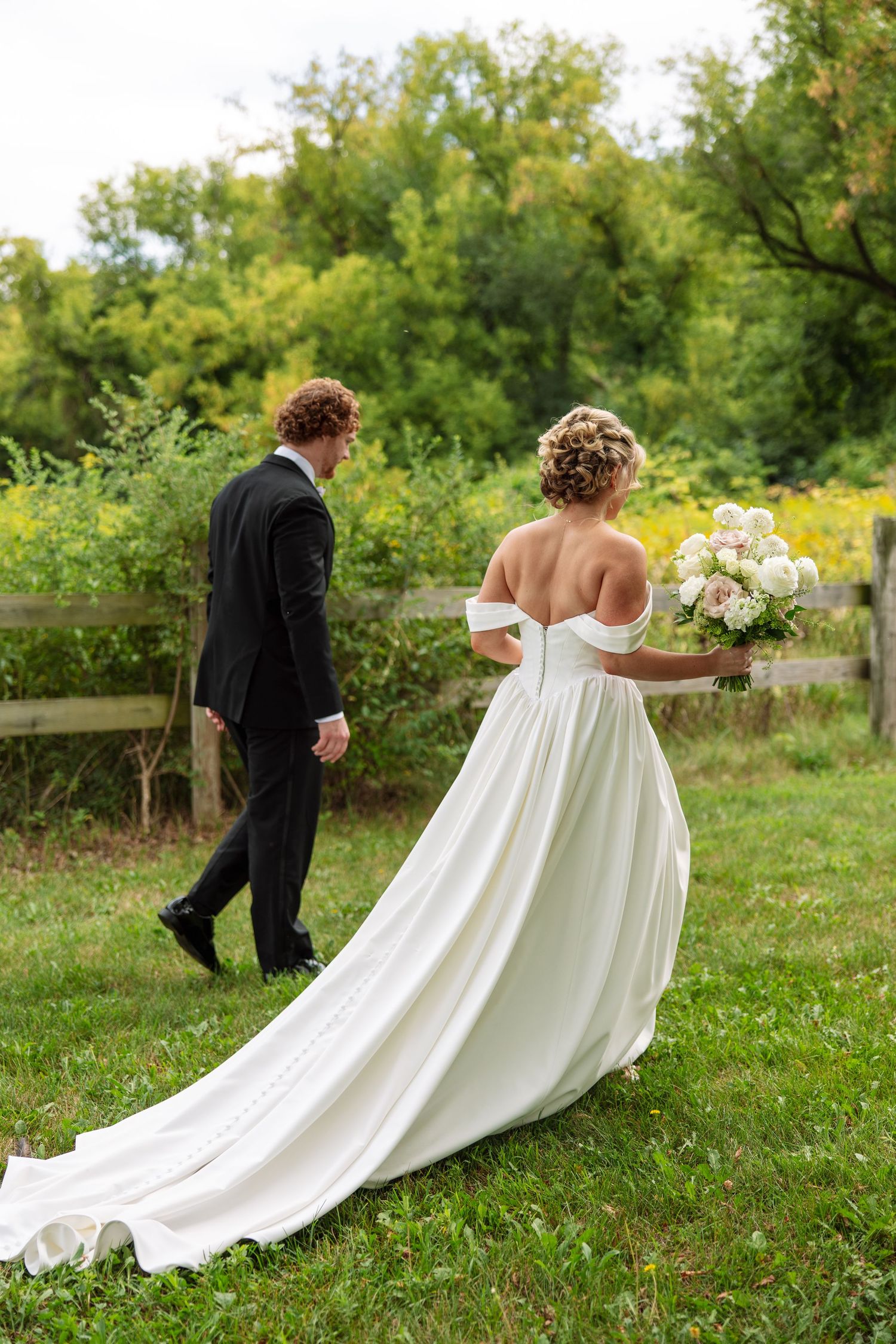 Bride in an off-the-shoulder satin gown with a long train holding a white bouquet walks beside the groom on a grassy path.