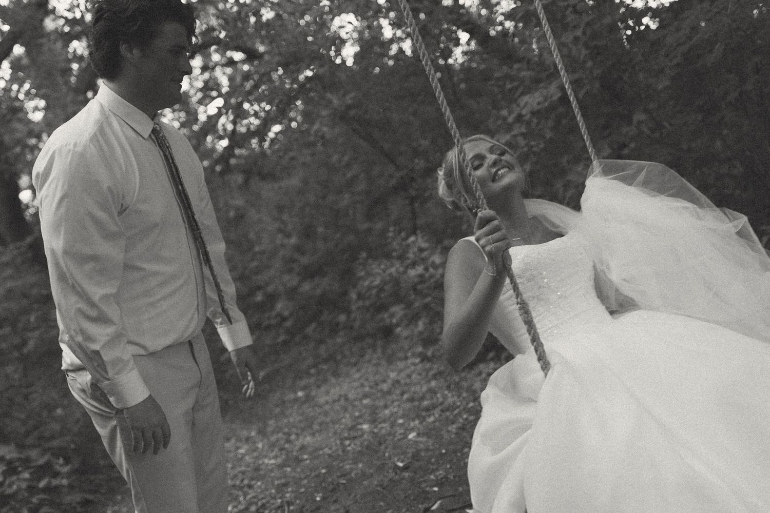 Black and white photo of a bride in a flowing gown laughing on a rope swing while the groom stands beside her in the woods.