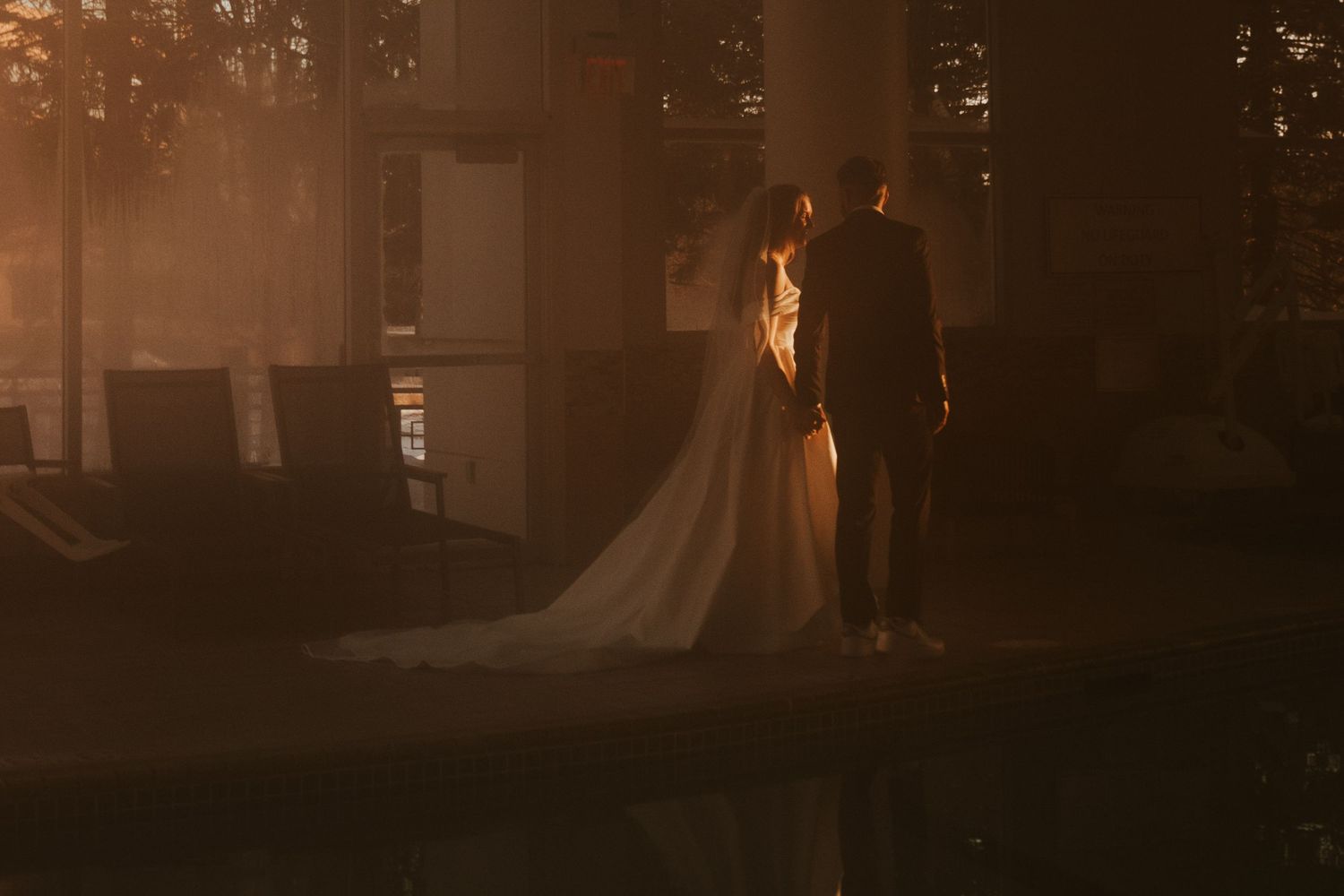 Bride in a flowing white gown and groom in a dark suit stand hand in hand in warm, golden light.
