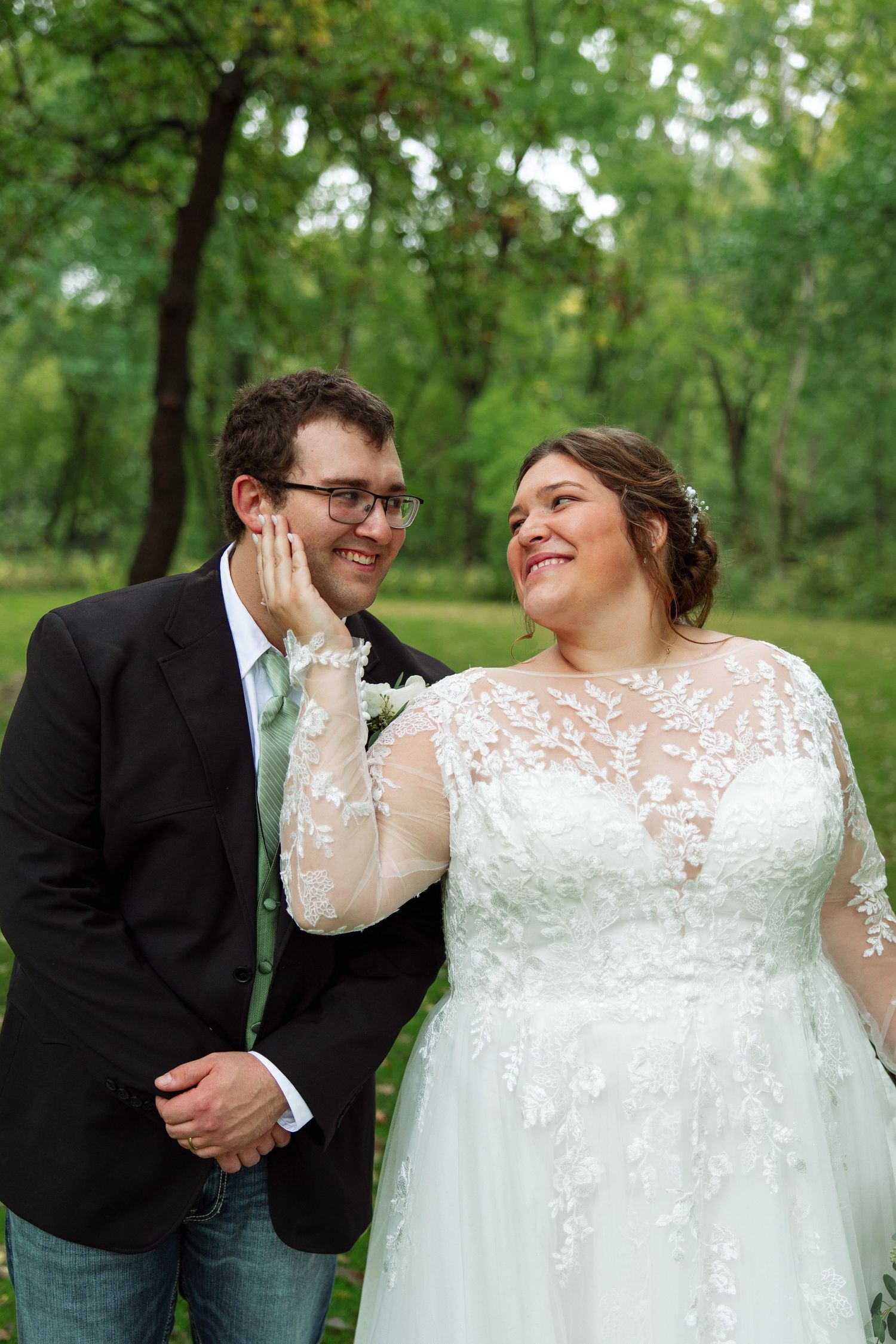 A smiling bride in a lace wedding gown and a groom in a dark jacket and glasses share a joyful moment in a green park.