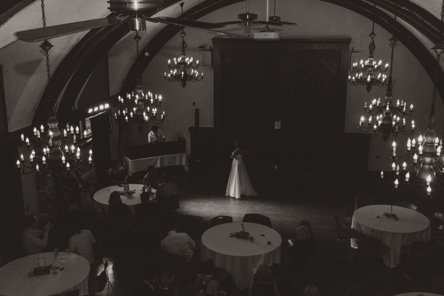 Black and white photo of a bride in a gown standing alone on a dance floor beneath chandeliers in an elegant wedding reception hall.