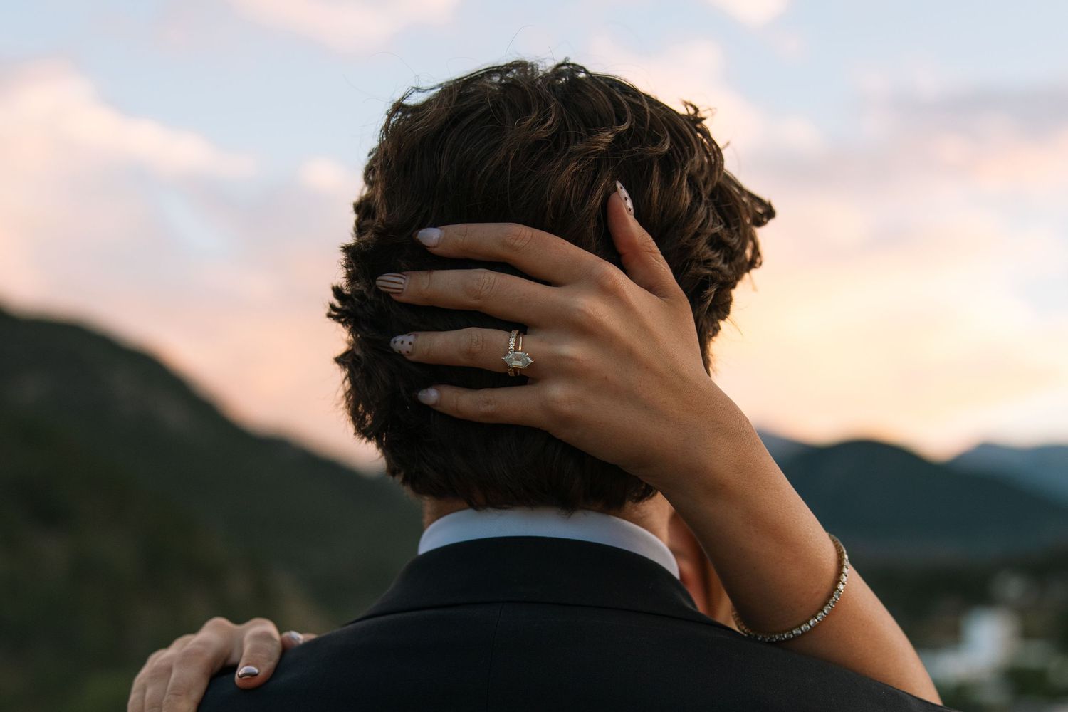 Close-up of a bride's hand with a diamond ring resting on her groom's head at sunset with mountains in the distance.