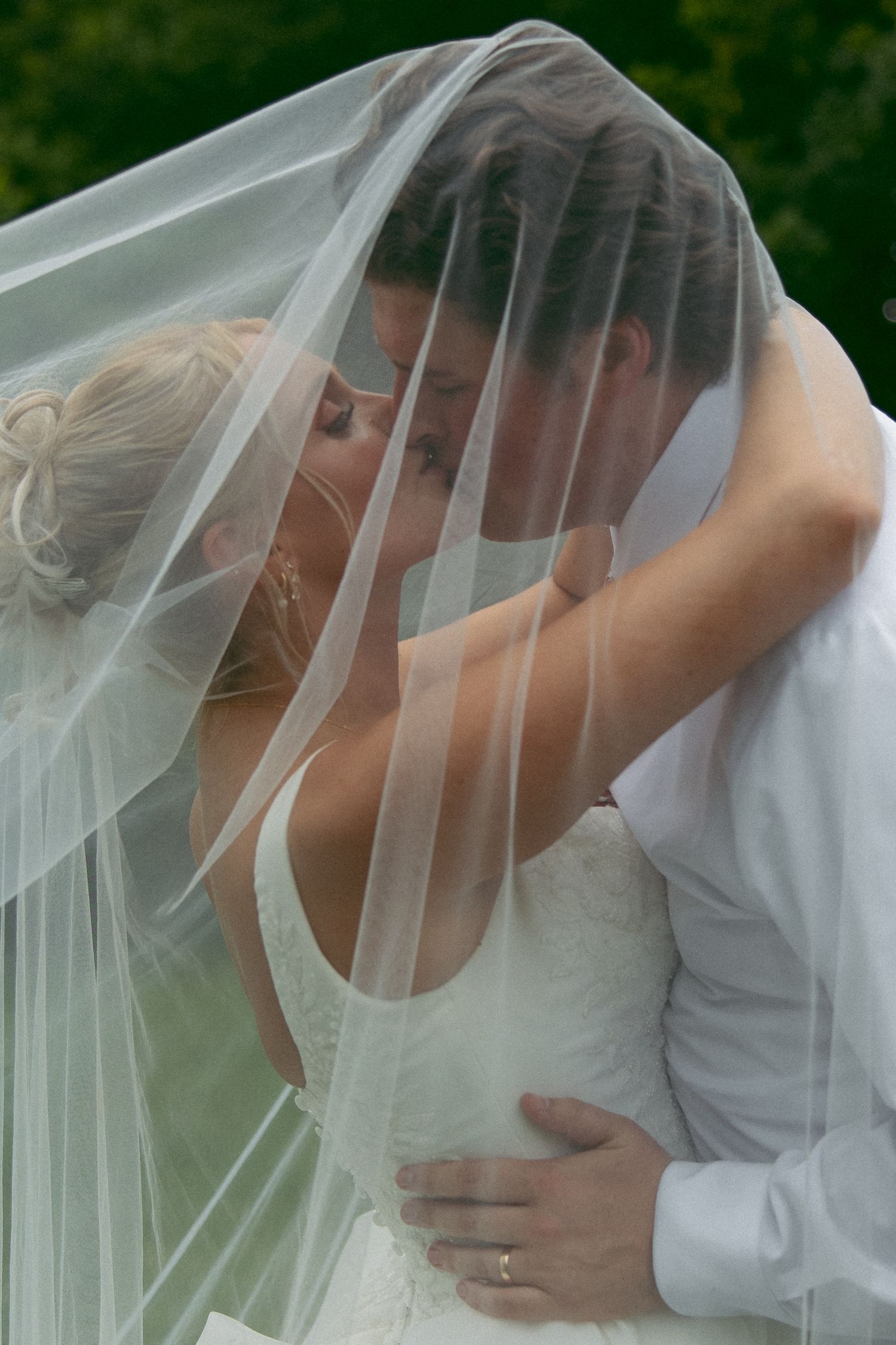 Bride and groom kiss beneath a sheer wedding veil in a tender embrace, with green foliage softly blurred behind.