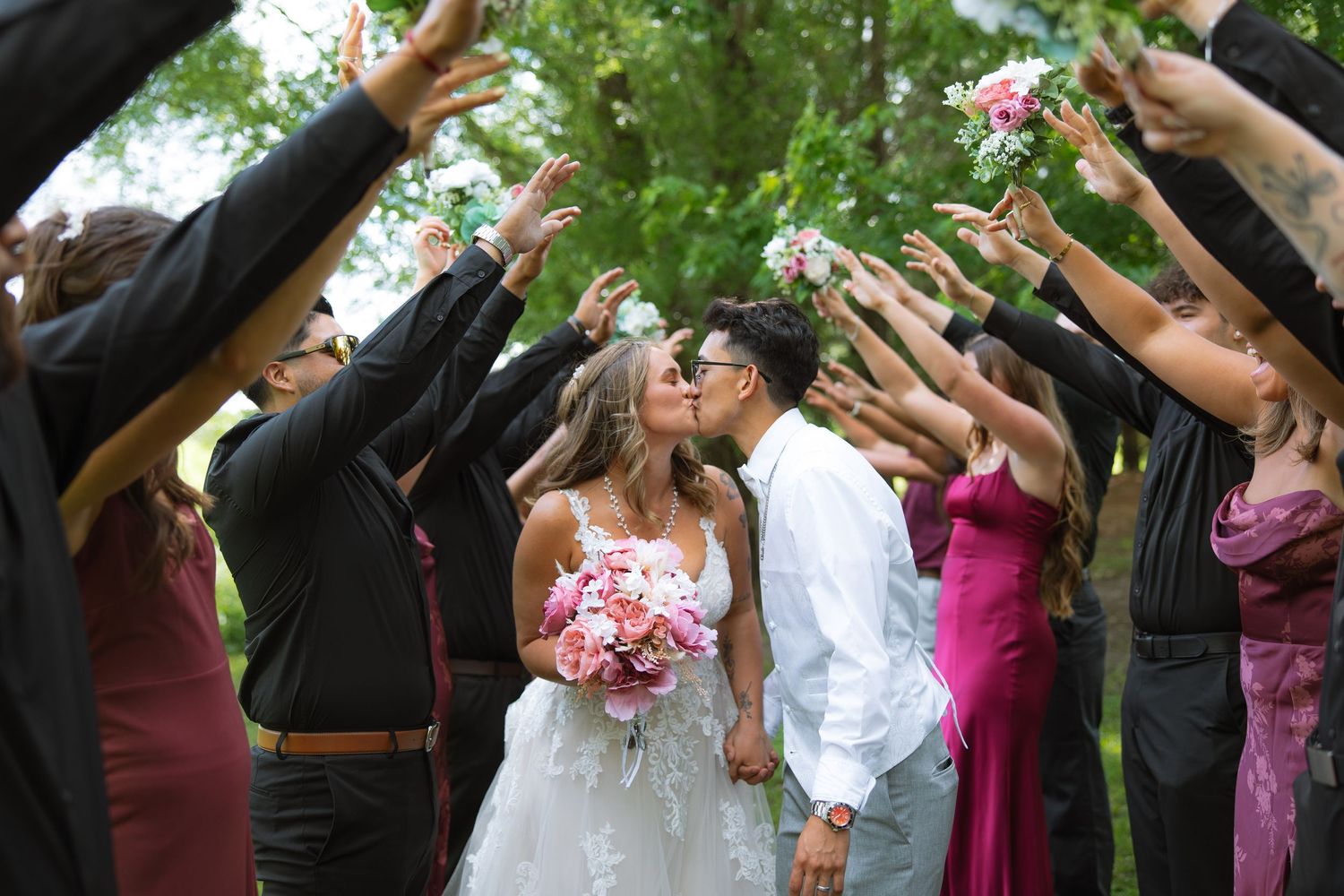 Bride and groom share a kiss as guests form an arch with raised hands, with the bride holding a pink and white bouquet.