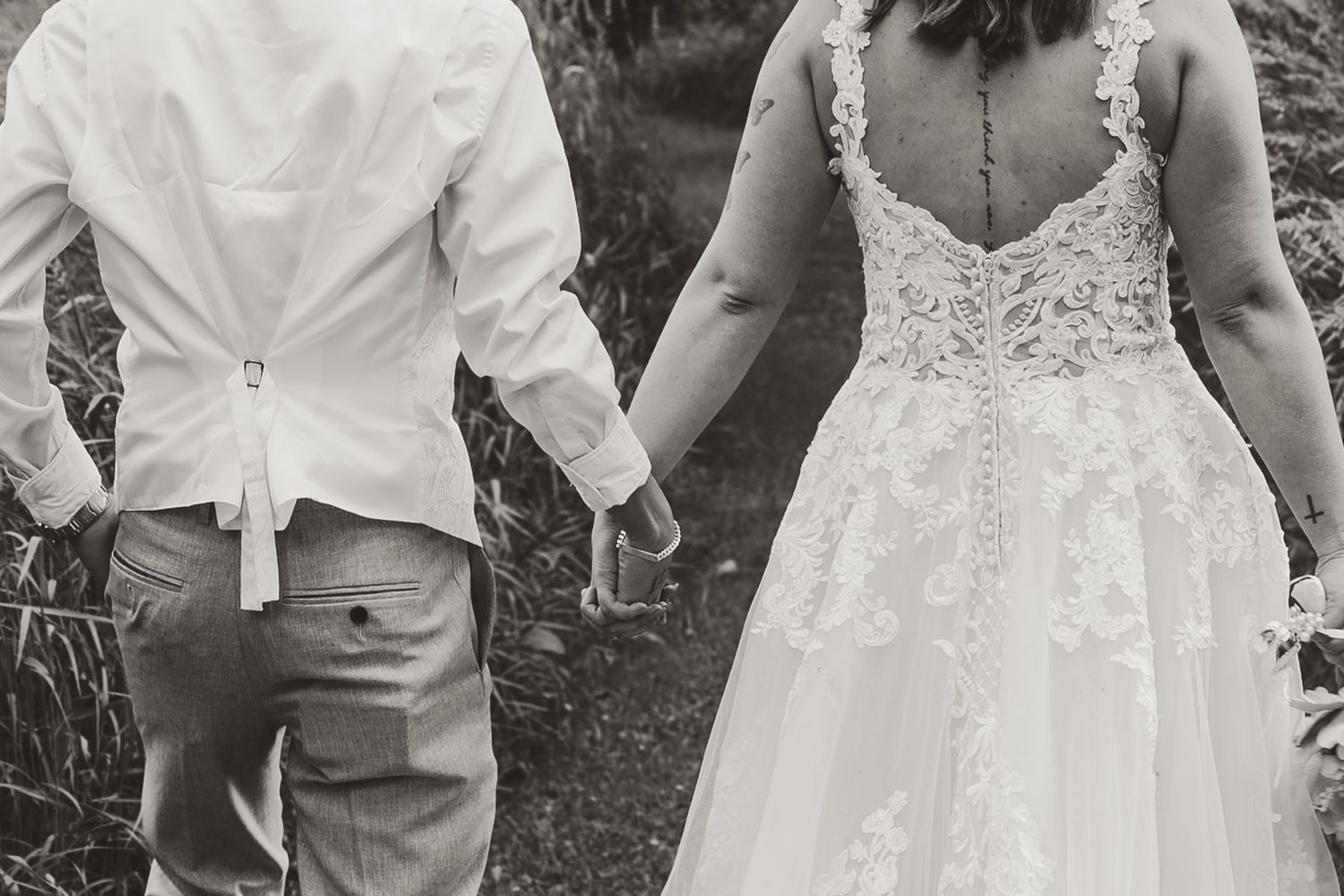 Black and white rear view of a bride in a lace-back wedding gown holding hands with her groom in a vest while walking outdoors.