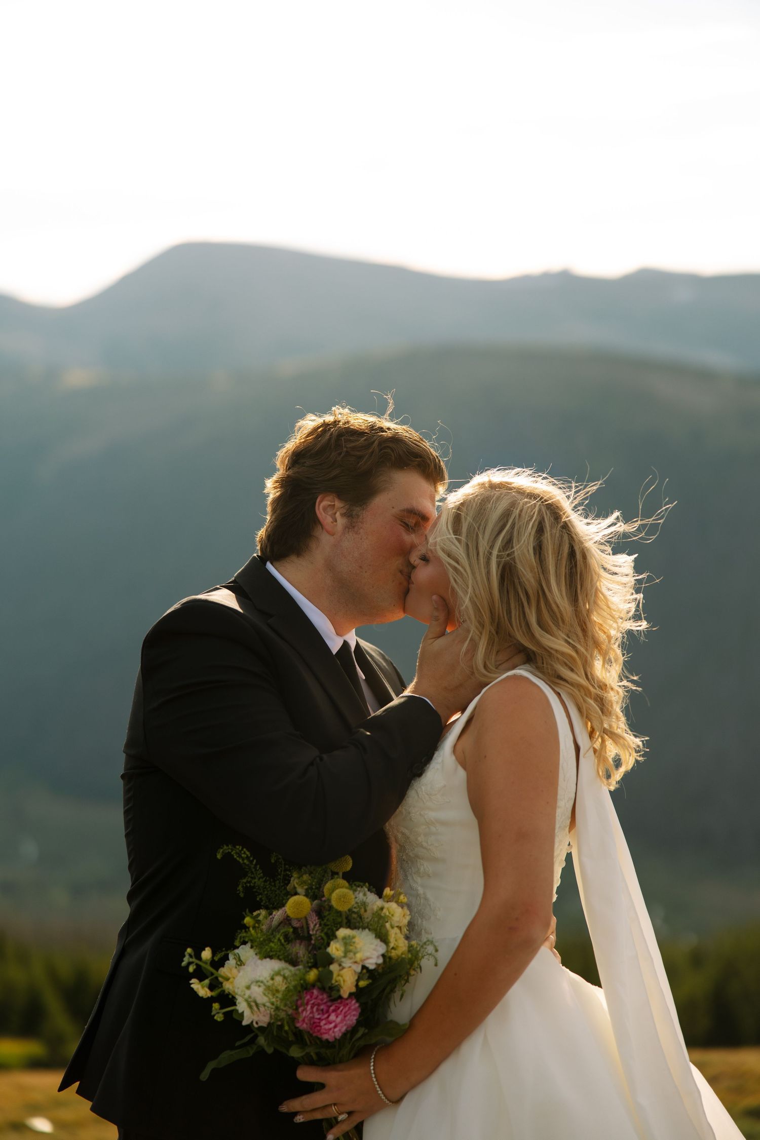 A bride and groom share a kiss outdoors with a wildflower bouquet, set against sunlit mountains at a scenic mountain wedding.