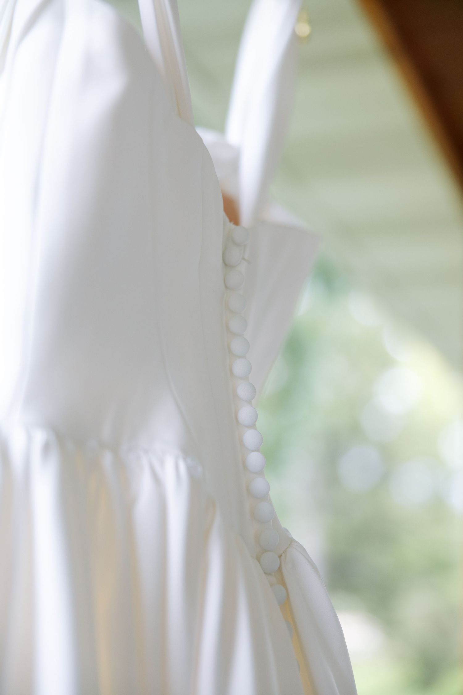 White satin wedding dress with a row of small covered buttons down the back, hanging against a softly blurred green background.