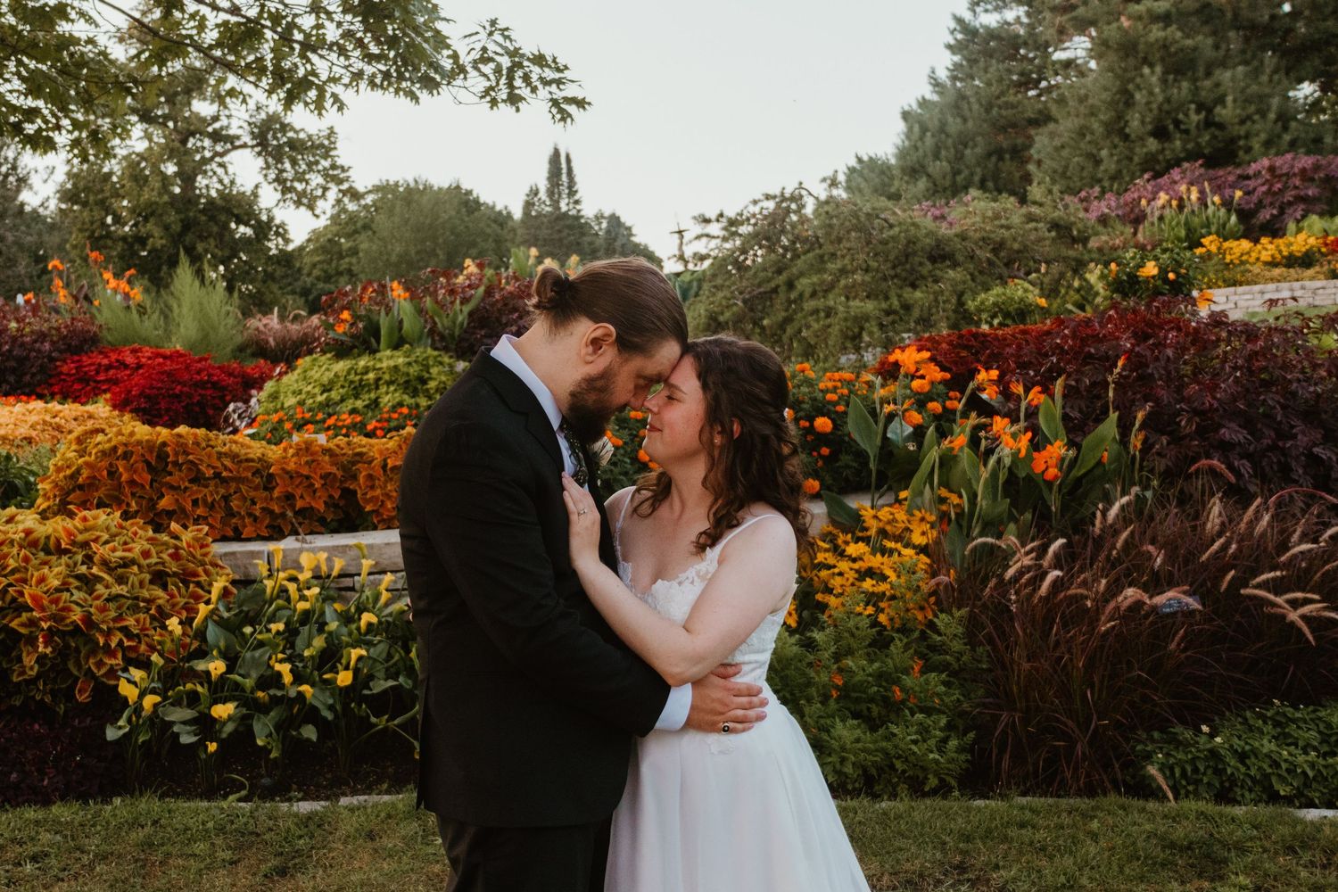 Bride in white gown and groom in dark suit embrace in a lush garden surrounded by colorful orange and red flowers.