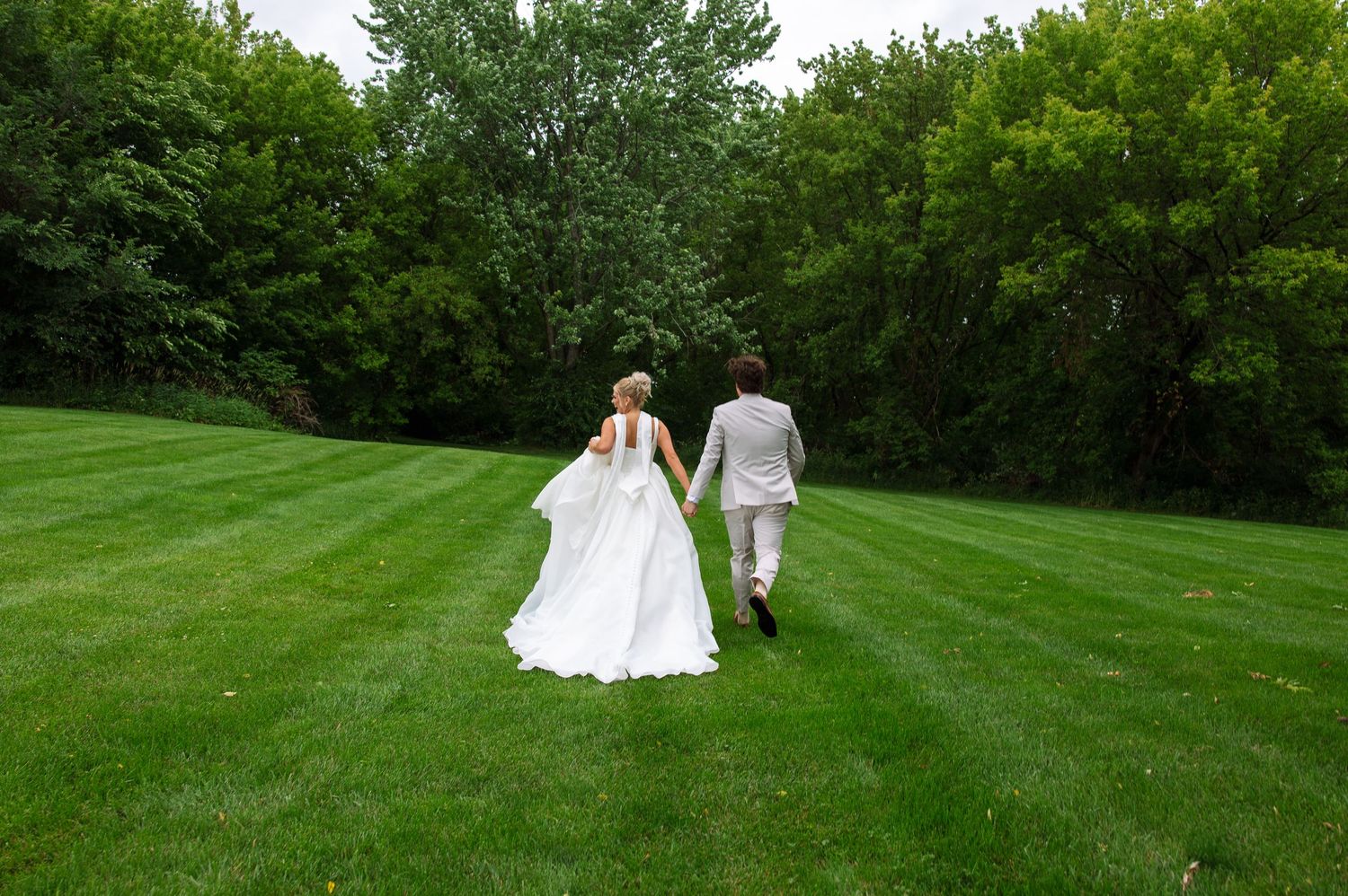 Bride in a flowing white gown and groom in a light suit hold hands while walking across a freshly mowed green lawn toward a tree line.