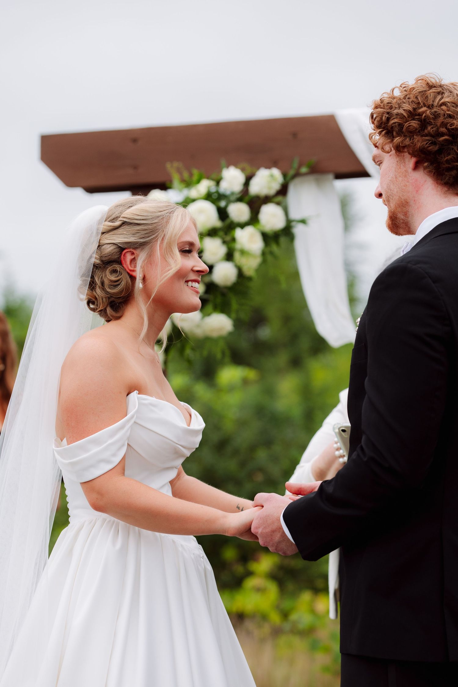 Smiling bride in a veil and off-the-shoulder gown holds hands with the groom beneath a floral wedding arch during the ceremony.
