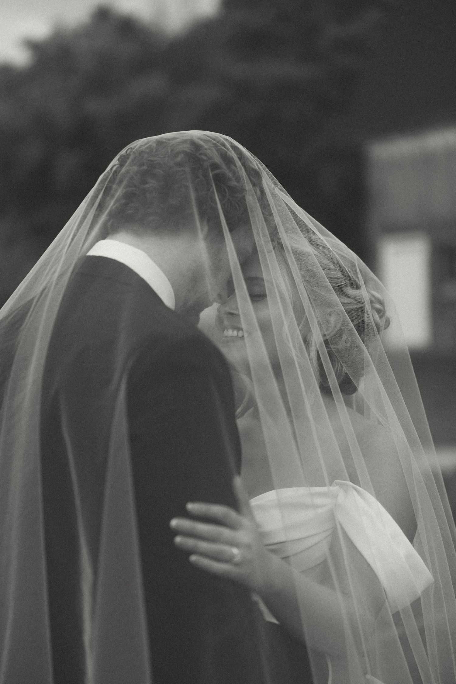 Black and white photo of a couple embracing and smiling beneath a sheer wedding veil outdoors.