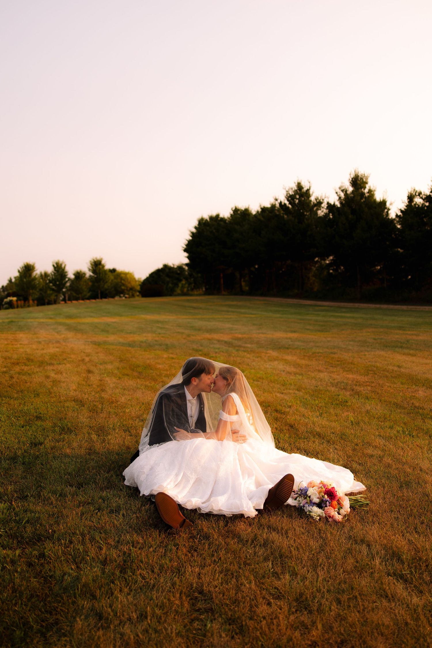 Bride and groom share an intimate moment seated on a grassy field at sunset, with a flowing veil, white gown and bouquet nearby.