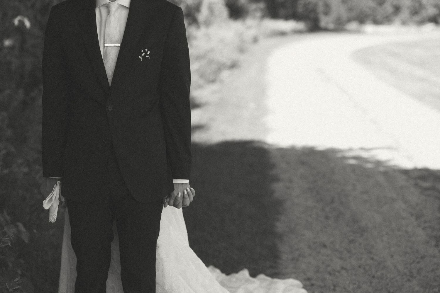 Black and white photo of a groom in a dark suit with pocket square standing on a country road holding his bride's veil.