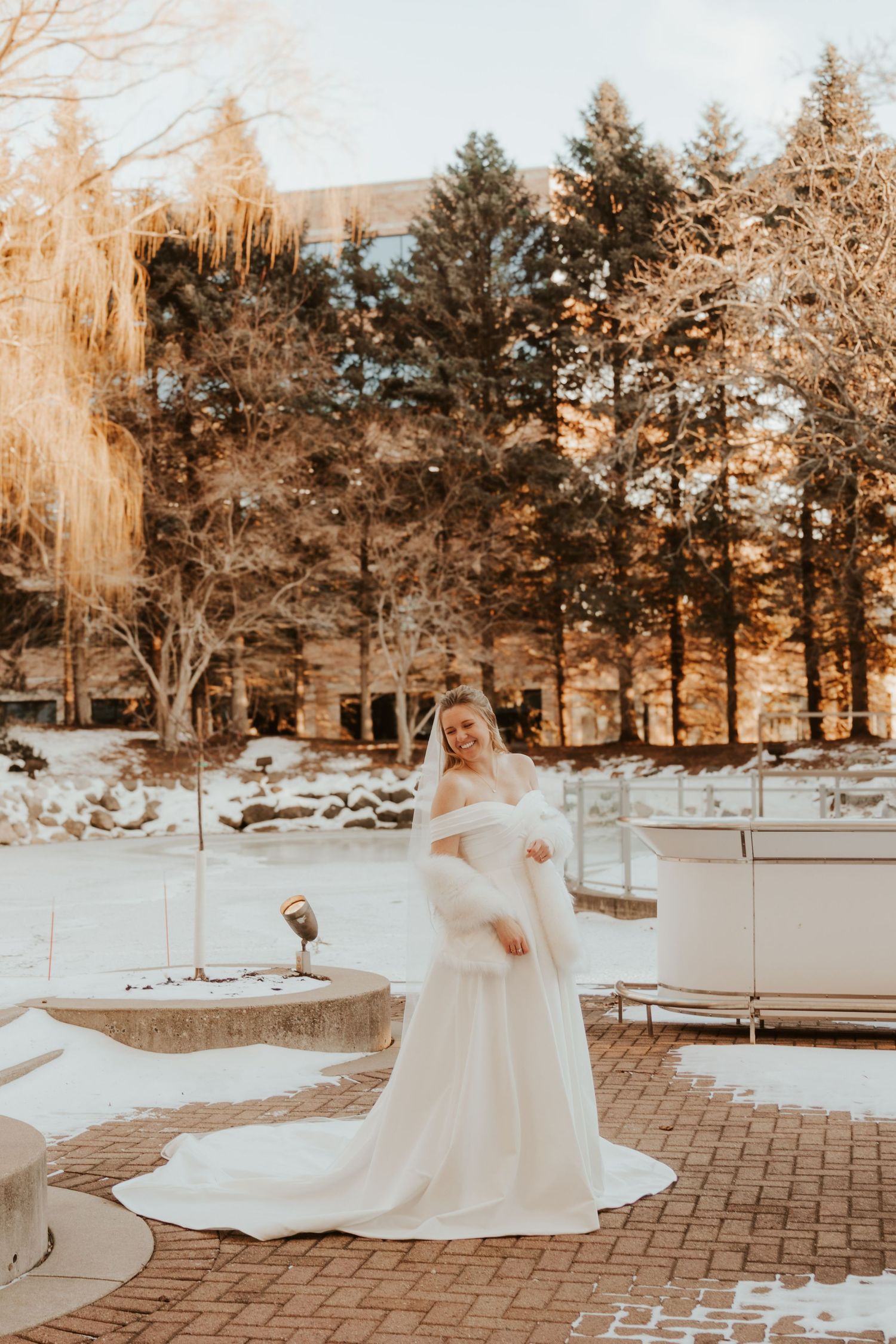 Bride in a white gown with a flowing train and fur shawl poses on a brick patio surrounded by snow and evergreen trees.