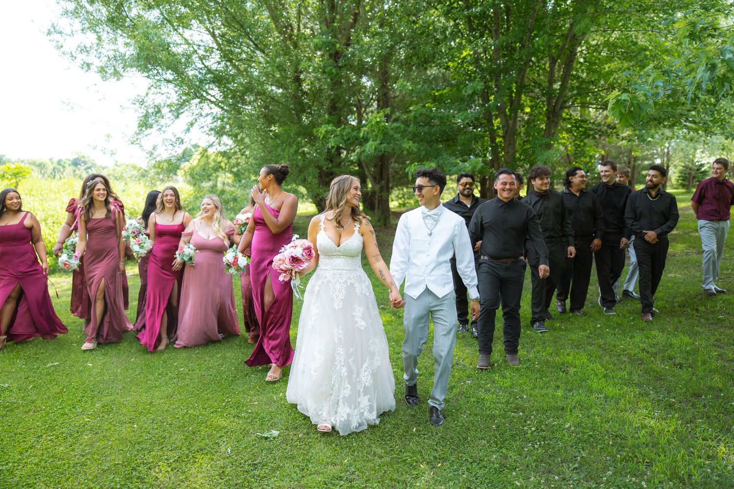 Bride and groom hold hands and walk across a sunny lawn, followed by smiling bridesmaids in mauve dresses and groomsmen in black.
