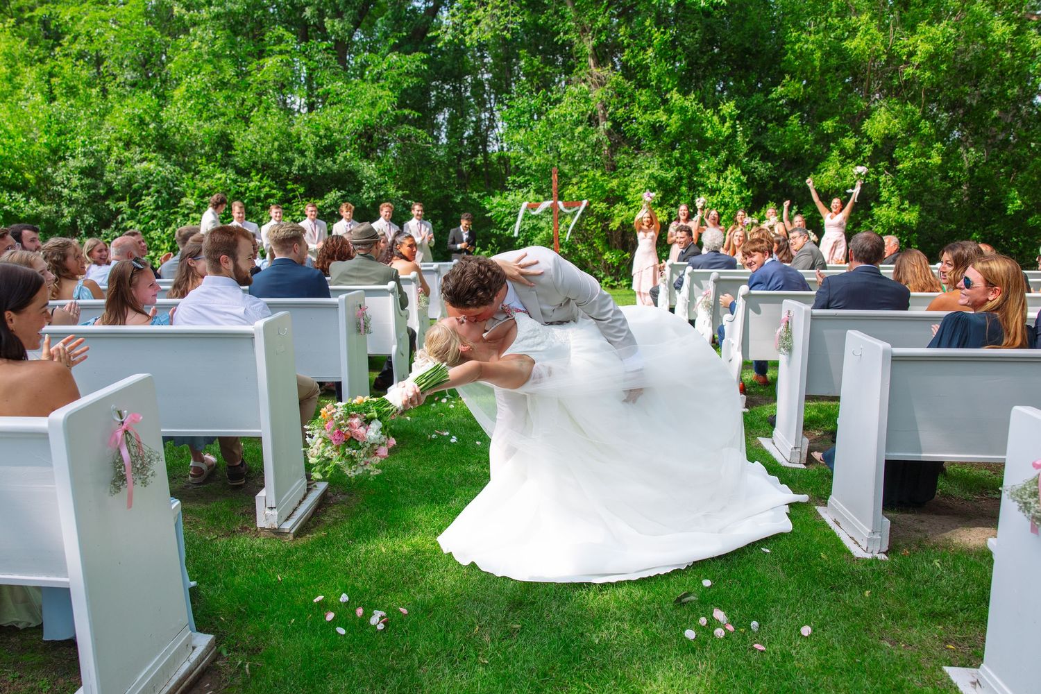 Groom dips and kisses the bride in the aisle of an outdoor wedding ceremony as guests cheer from white benches.