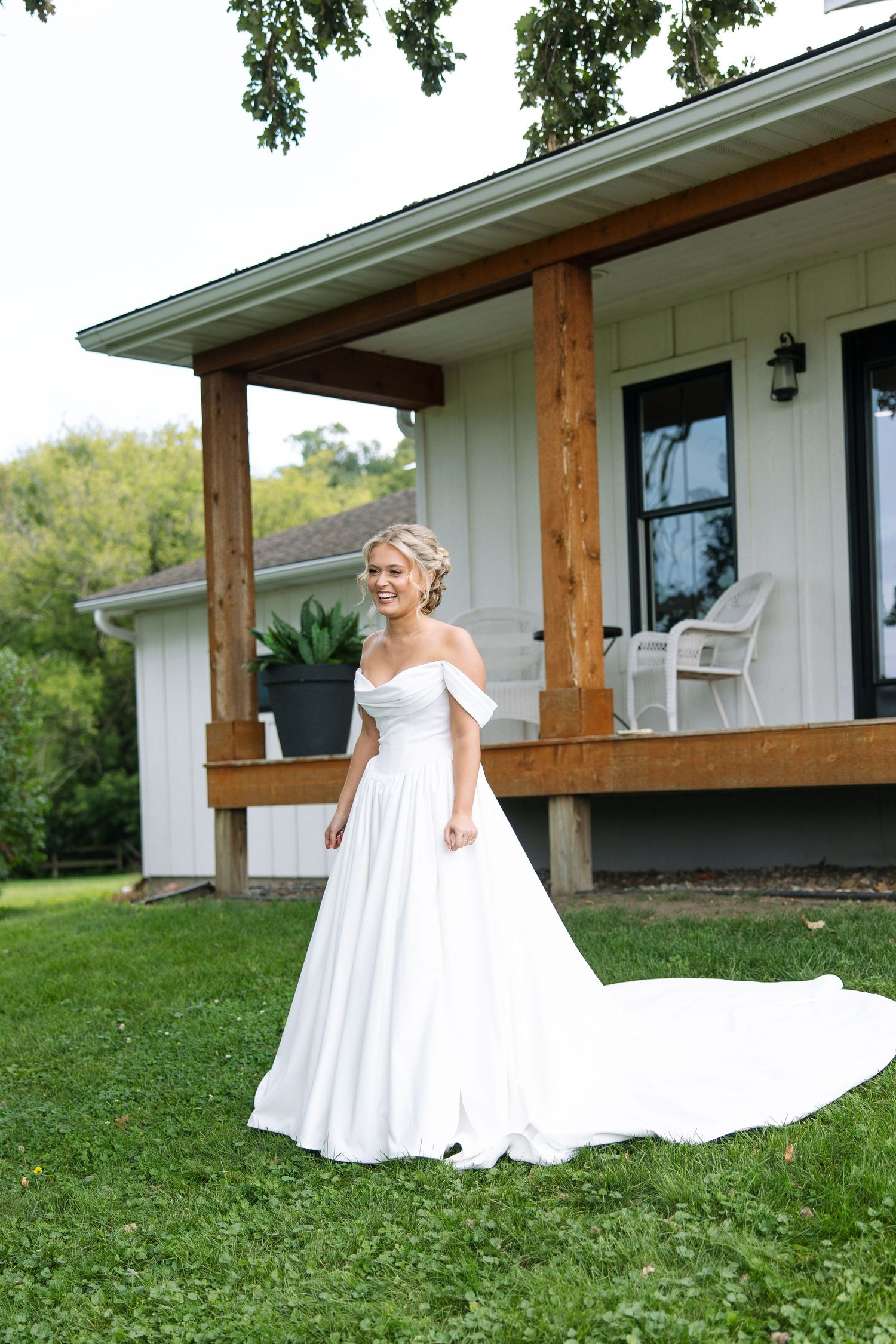 Bride in an off-the-shoulder white satin wedding gown with a long train stands on green grass before a rustic wood-beam porch.