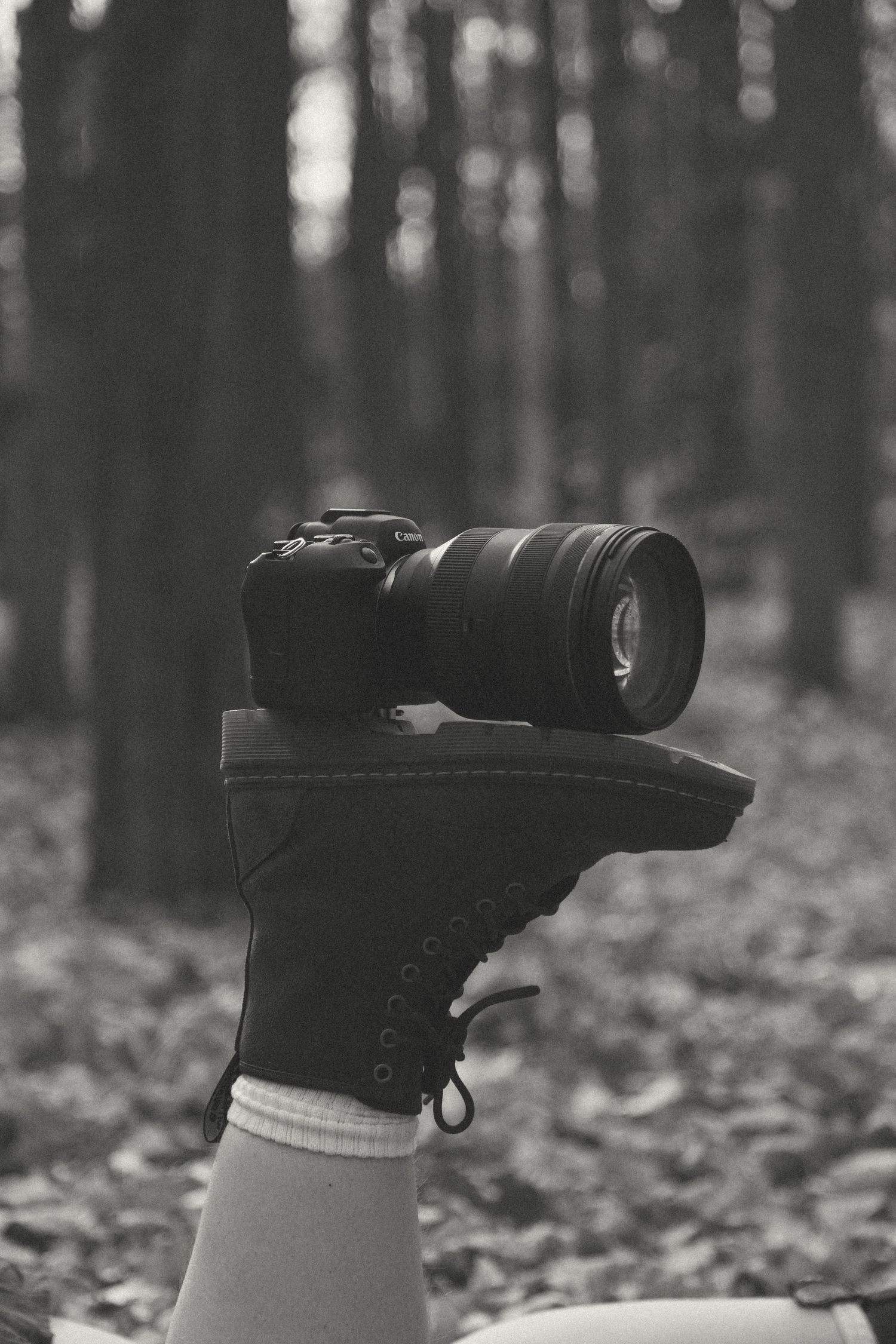 Black and white photo of a camera with a large lens balanced on the sole of a lace-up boot raised in a leaf-covered forest.