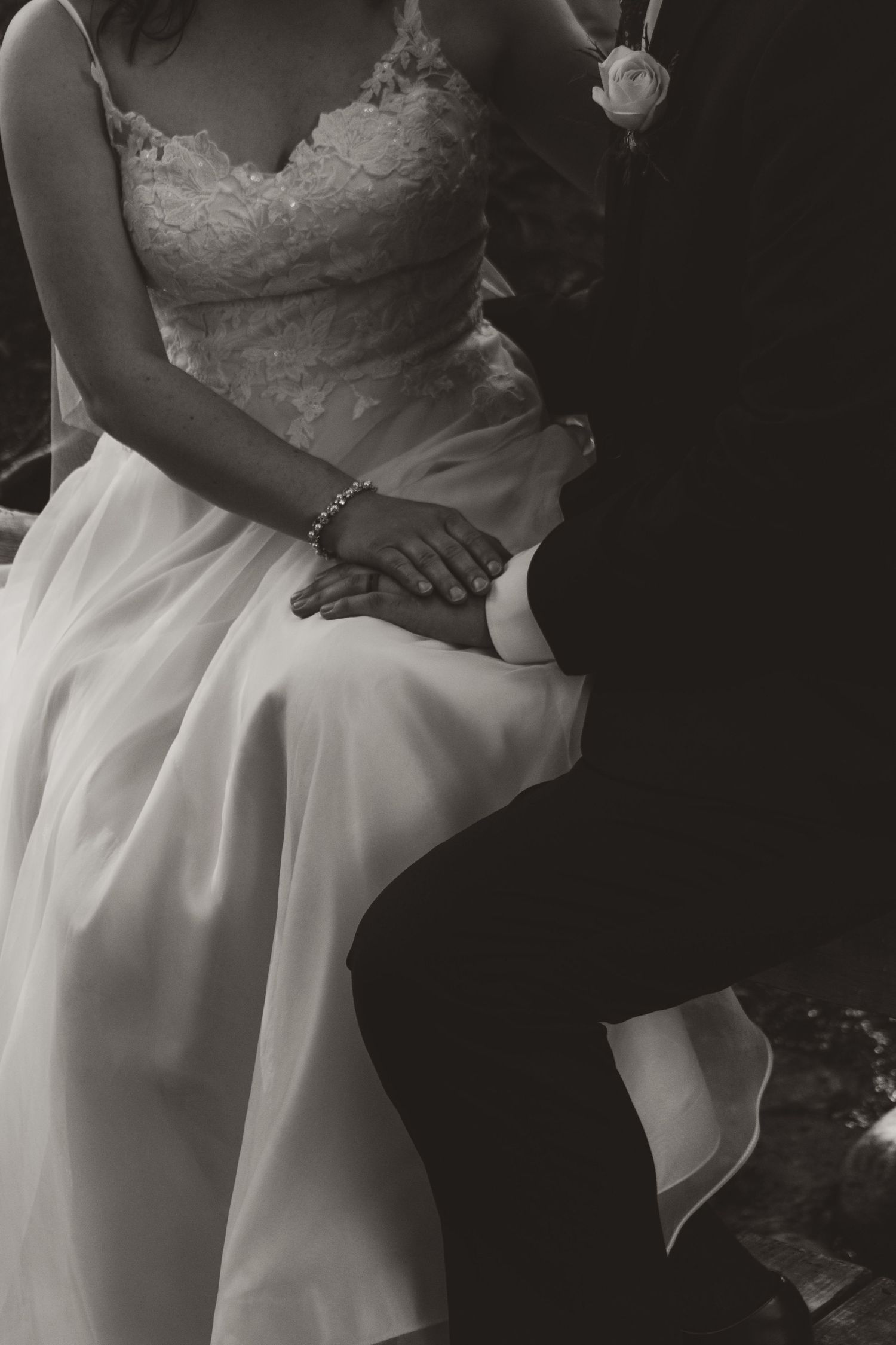Black and white photo of a bride in a lace wedding gown holding hands with a groom in a dark suit, a boutonniere on his lapel.