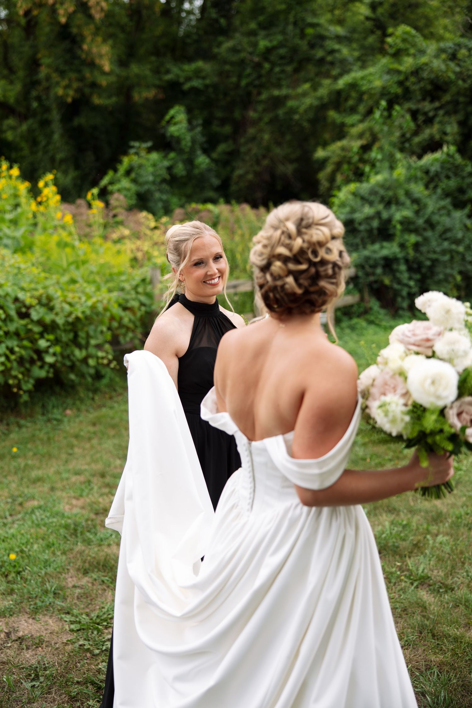 Bride in an off-the-shoulder satin gown holding a pink and white bouquet smiles at a bridesmaid in a black halter dress in a garden.