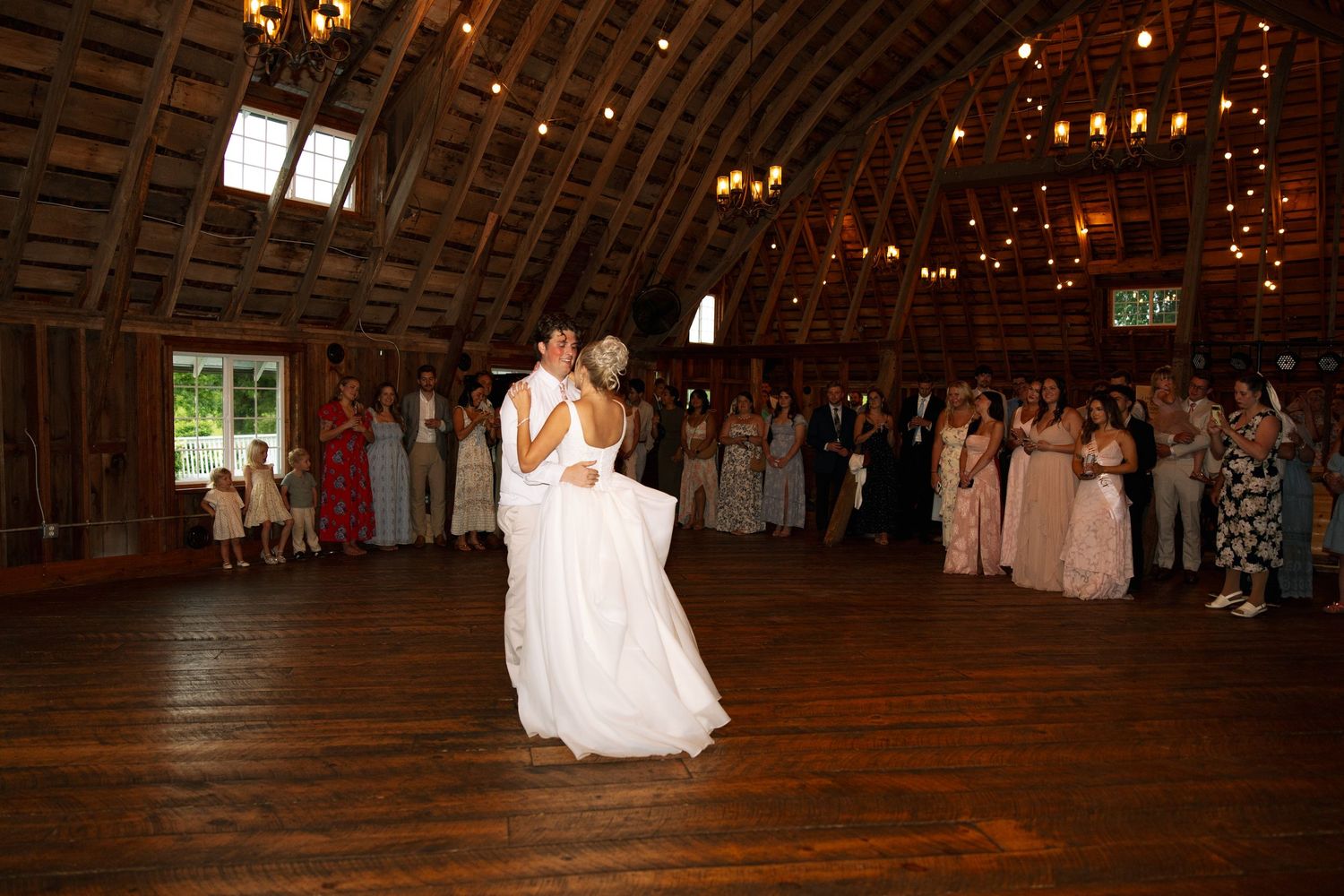 Bride and groom share their first dance on a wooden floor in a rustic barn venue while wedding guests watch.