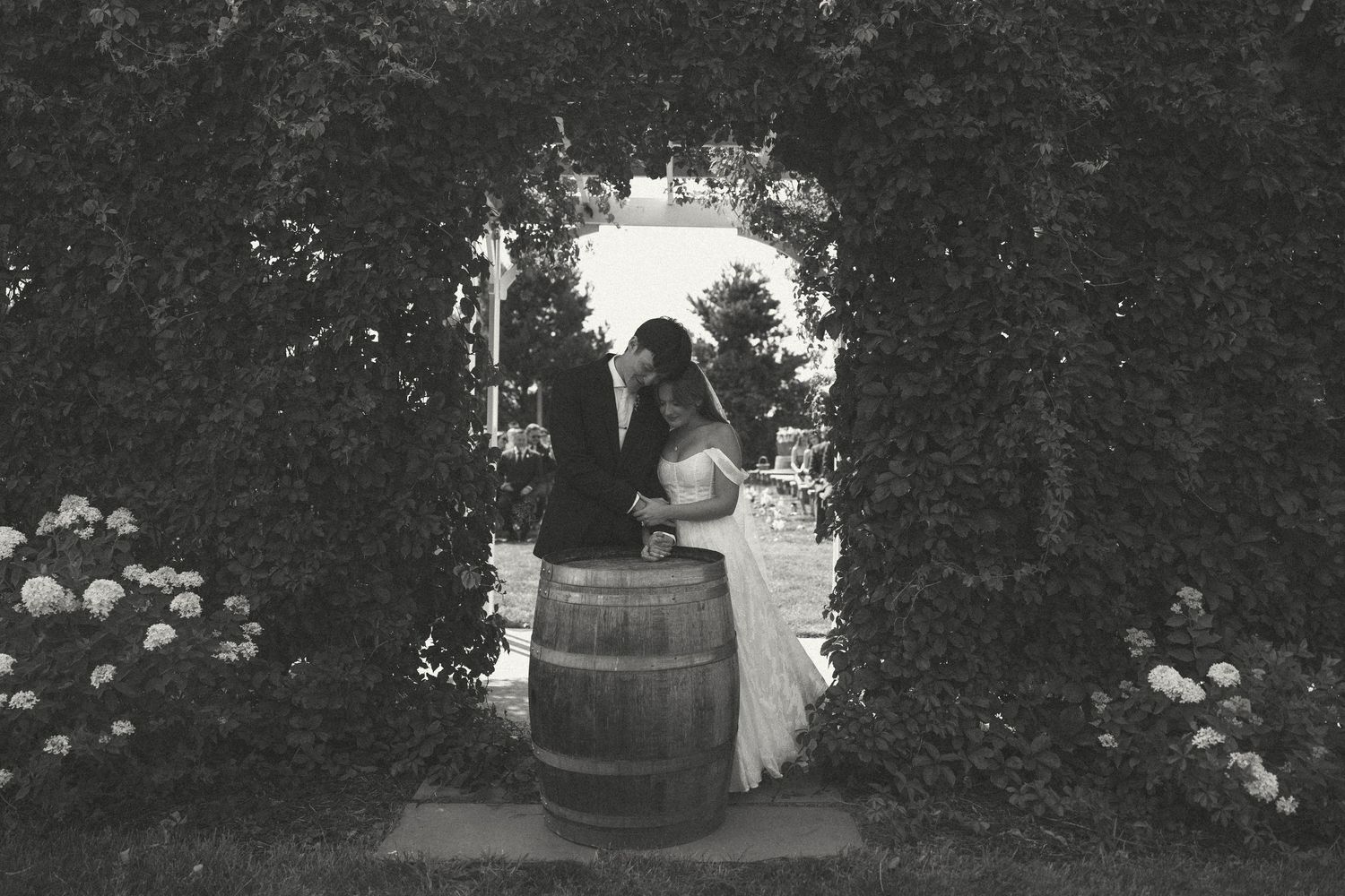 Black and white photo of a couple embracing beside a wooden barrel beneath a leafy garden archway with blooming hydrangeas.