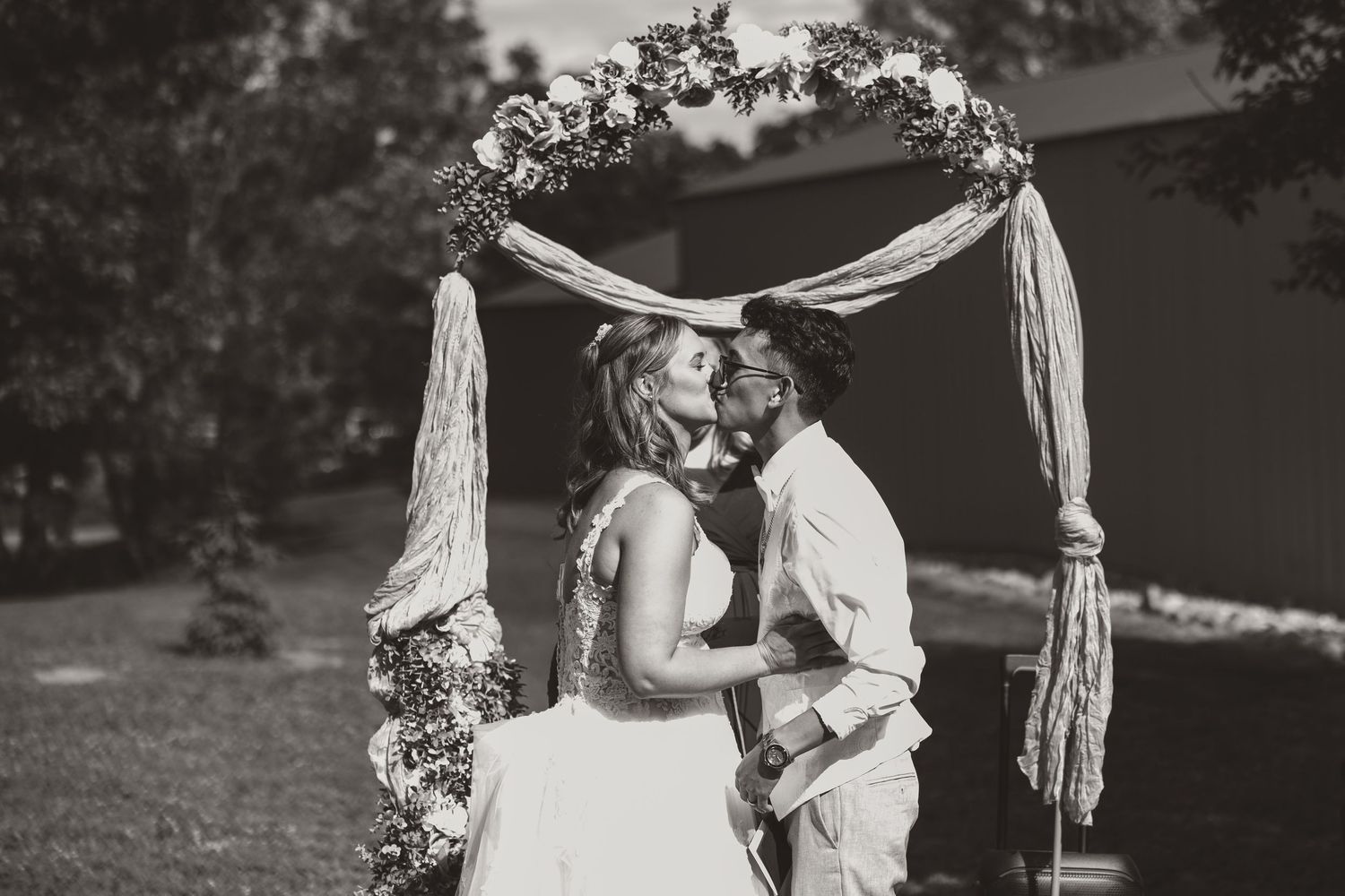 Bride and groom kiss beneath a rustic wedding arch draped with fabric and flowers in a black and white outdoor ceremony photo.