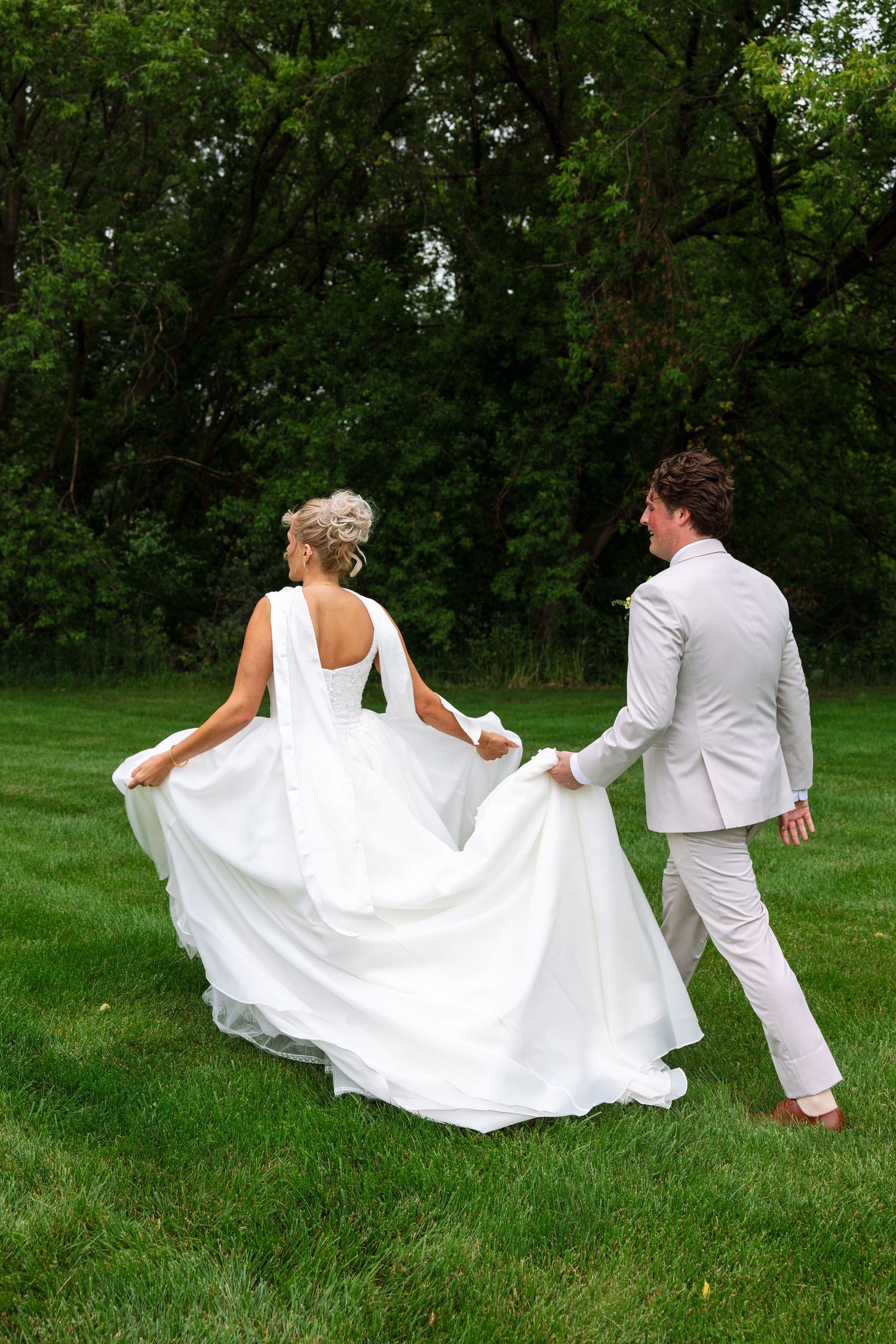 Bride lifts her long white gown while walking across a lush green lawn beside the groom in a light gray suit.