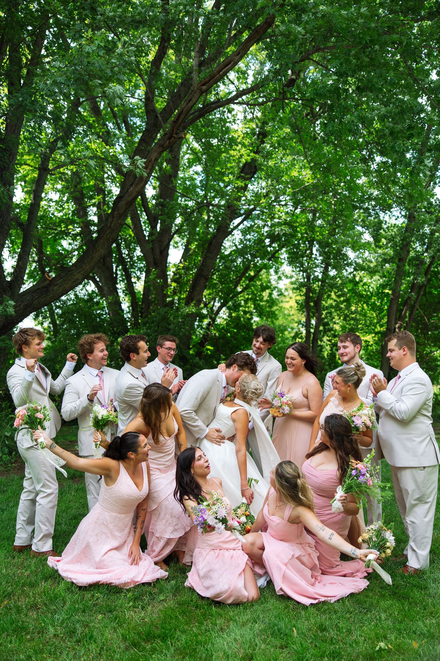 Bride in white gown laughs with bridesmaids in blush pink dresses and groomsmen in light suits on a green lawn beneath tall trees.