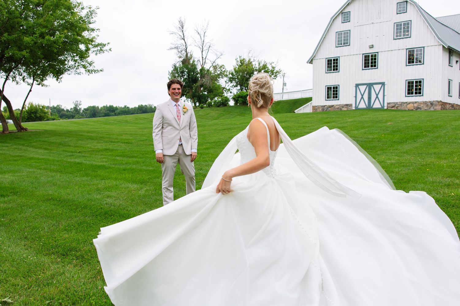 Bride twirls her flowing white gown on a green lawn as the groom in a tan suit watches near a white barn venue.