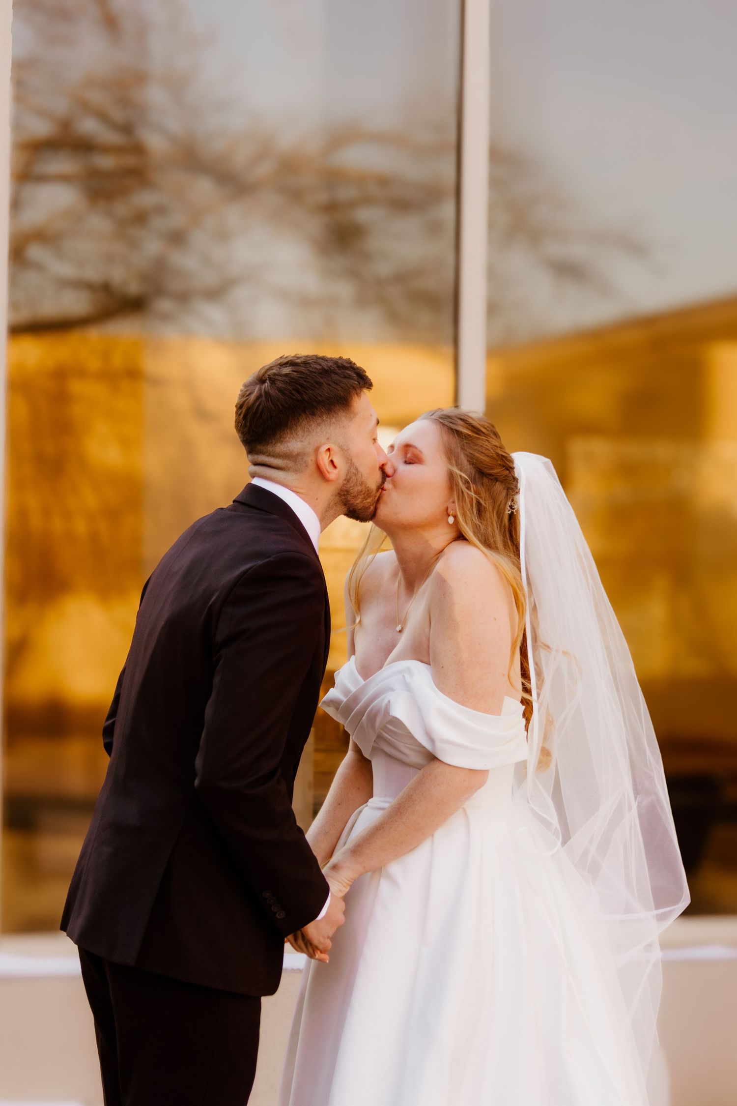 Bride in an off-the-shoulder gown and groom in a black suit share a kiss in front of a golden reflective window.