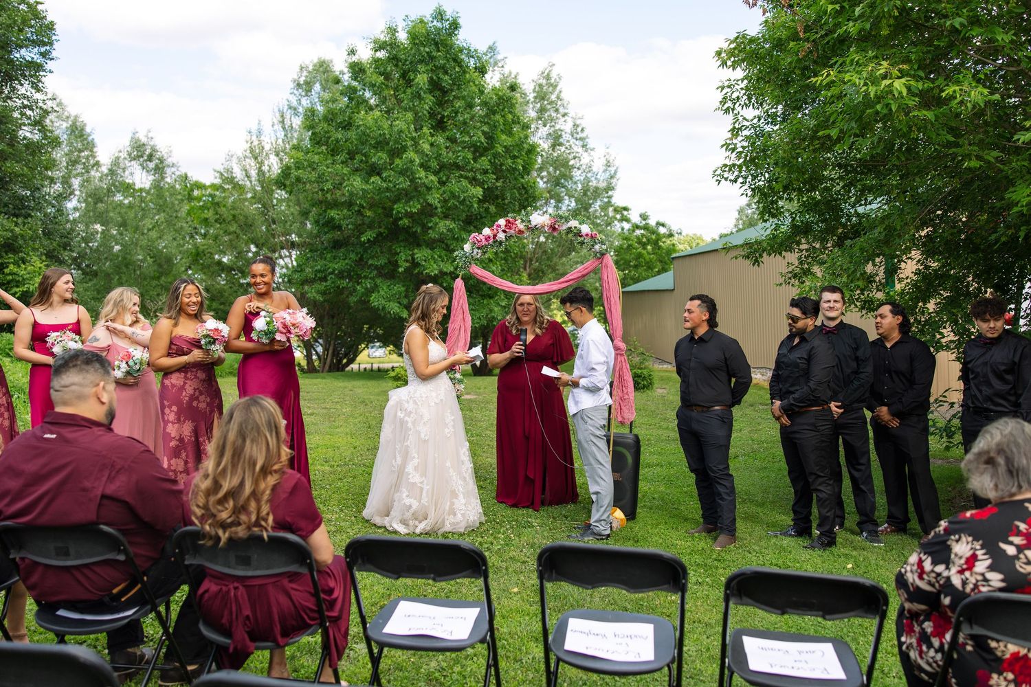 Outdoor wedding ceremony on a lawn with bridesmaids in burgundy dresses, groomsmen in black and guests seated before a pink floral arch.