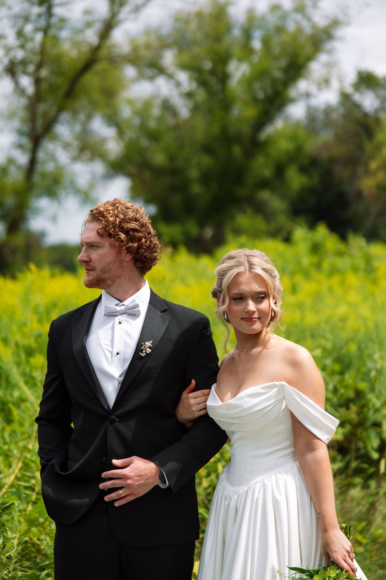 Bride in an off-shoulder white wedding gown links arms with a groom in a black tuxedo and bow tie in a sunny yellow wildflower field.