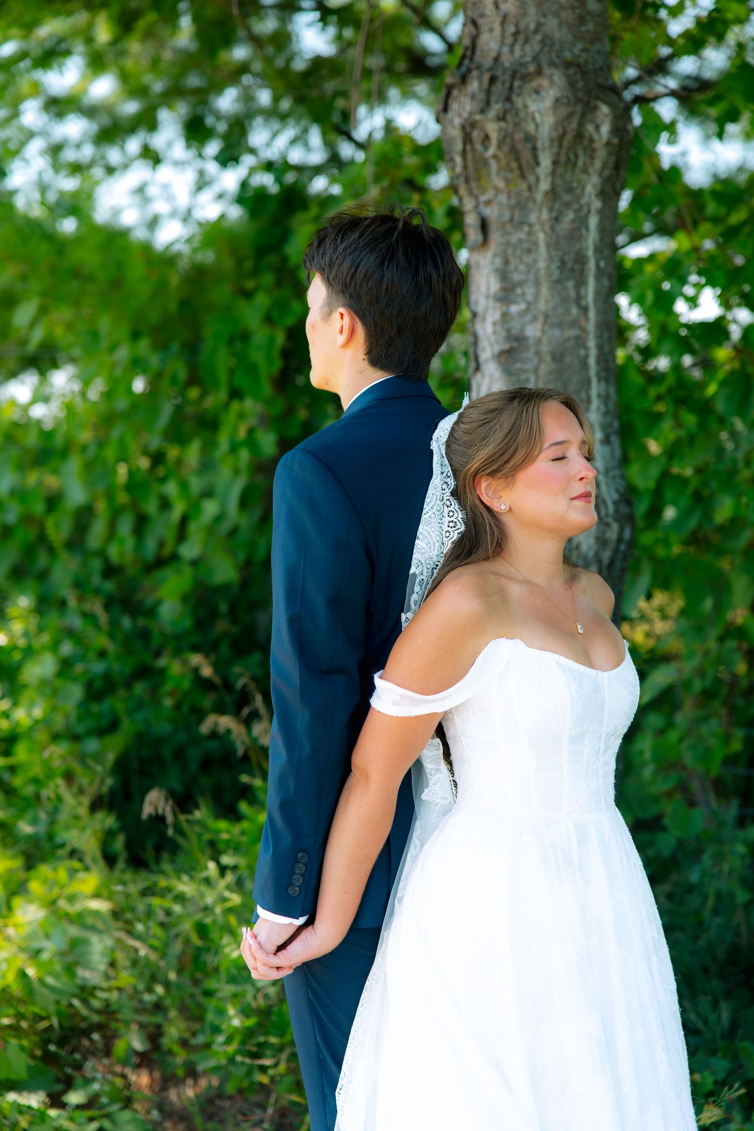 Bride in a white off-the-shoulder gown and lace veil stands back to back with a groom in a navy suit, holding hands beneath a tree.